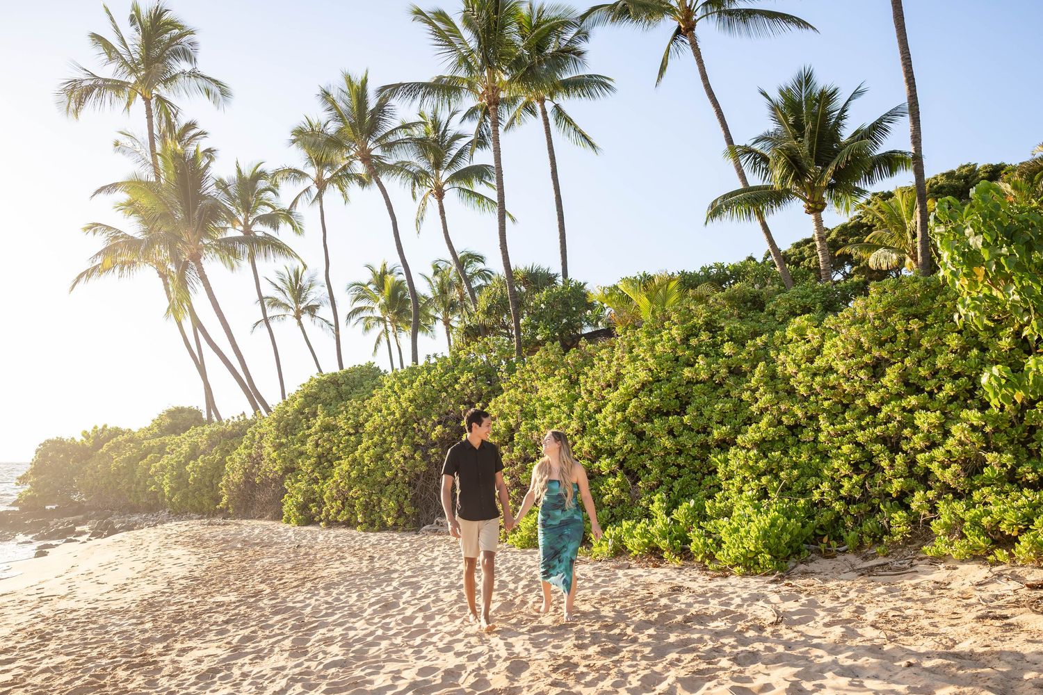Engaged couple walking hand in hand on a sandy Hawaiian beach surrounded by tall palm trees.