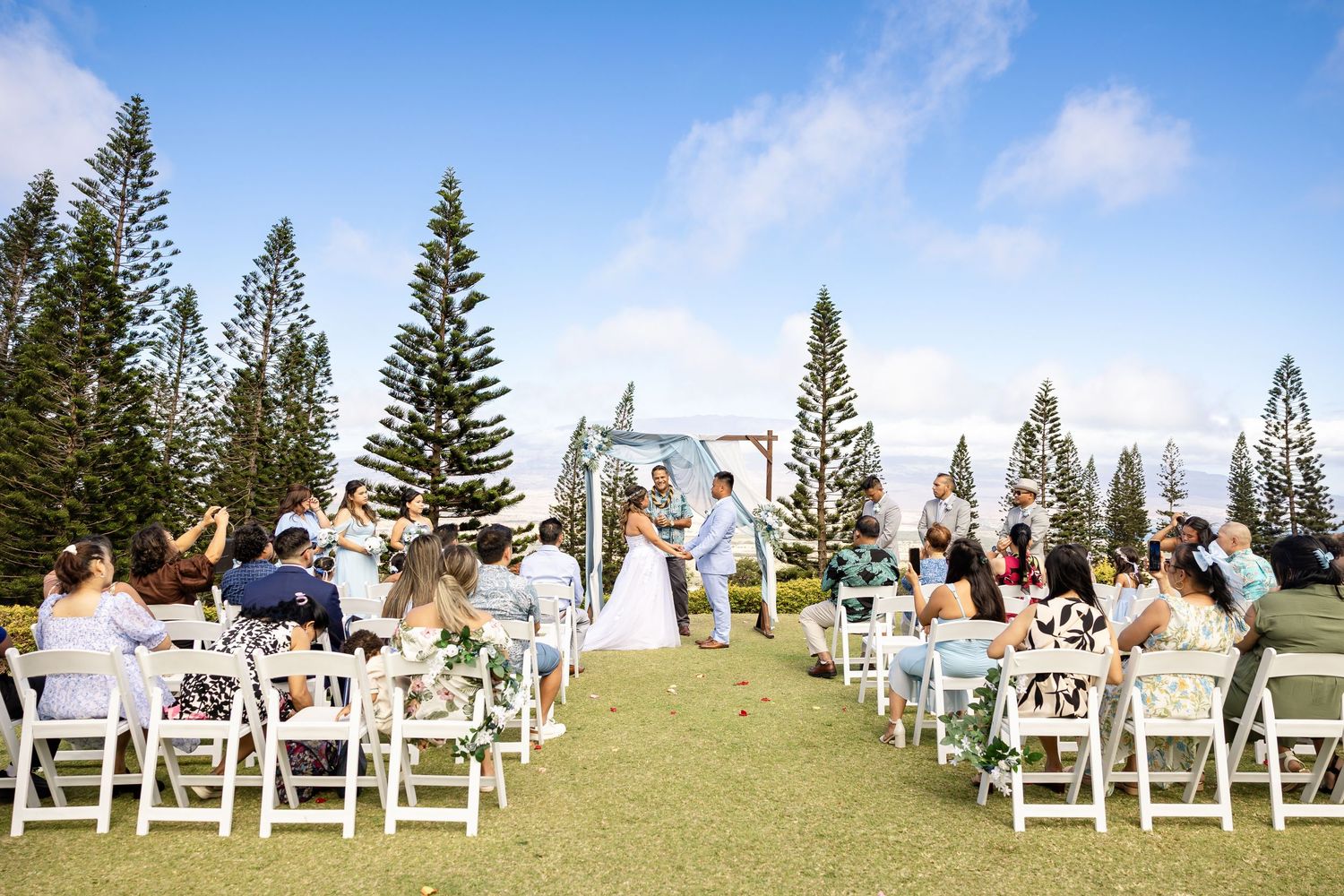 Outdoor wedding ceremony on a green lawn surrounded by tall pine trees in Hawaii.