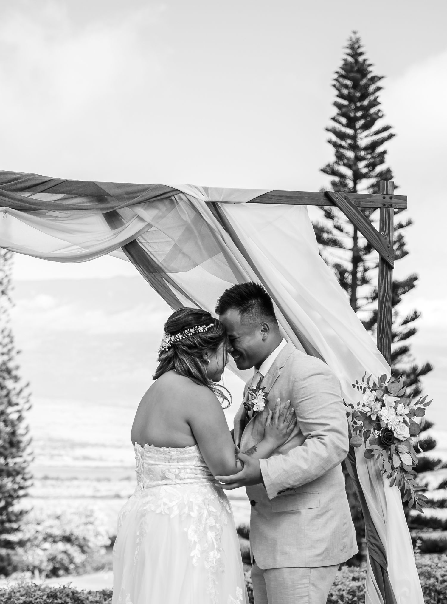 Bride and groom sharing intimate moment under draped wedding arch with pine tree backdrop in black and white.