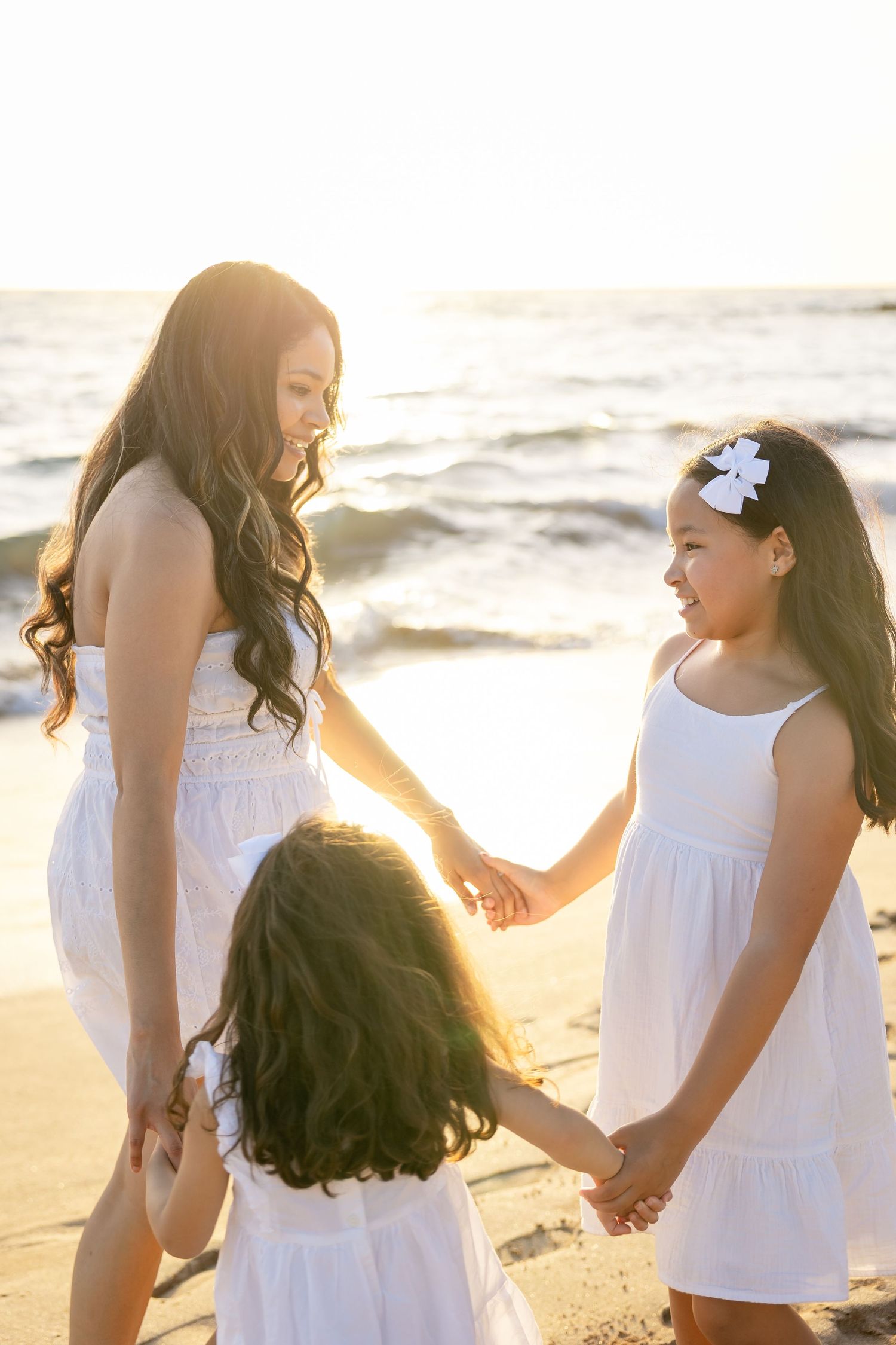Mother and two daughters in white dresses hold hands on a sunny beach at sunset.