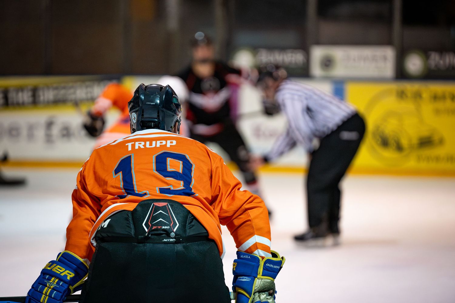 Player Trupp wearing jersey number 19 in orange uniform plays sledge hockey on ice rink.