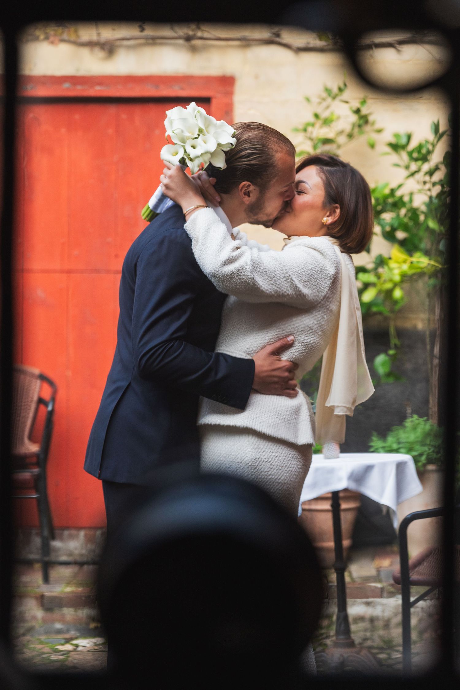 A couple sharing a romantic kiss, the bride holding white flowers, in front of a red door.