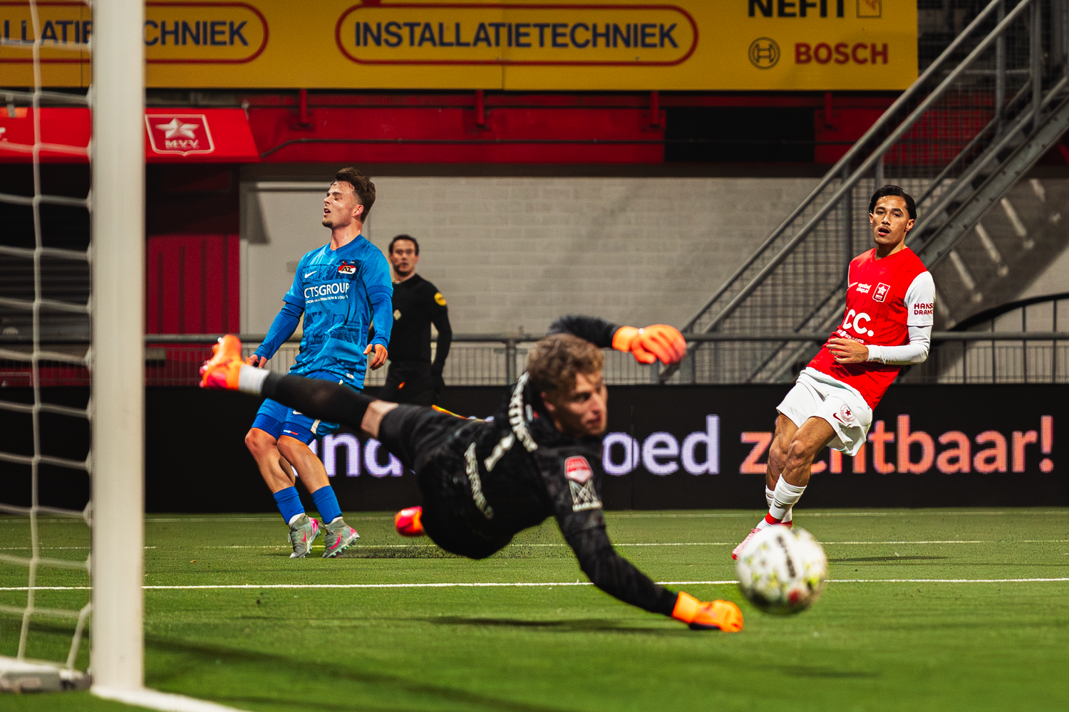 Soccer player in blue jersey takes a shot on goal while goalkeeper in black dives to make the save during match.