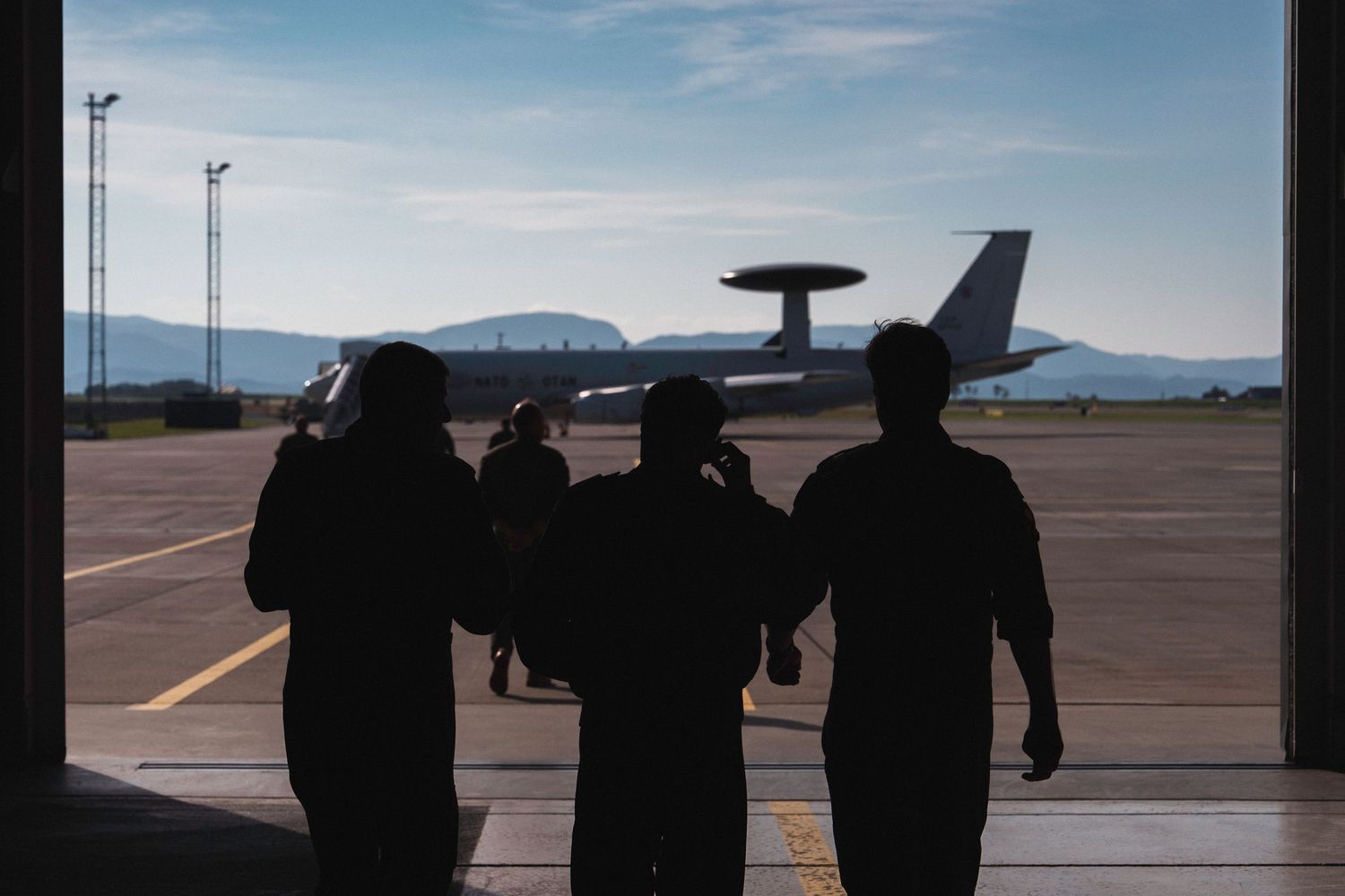 Silhouettes of E-3A Aircrew members walking to the aircraft for a mission. The plane is layered against a bright sky and mountain backdrop.