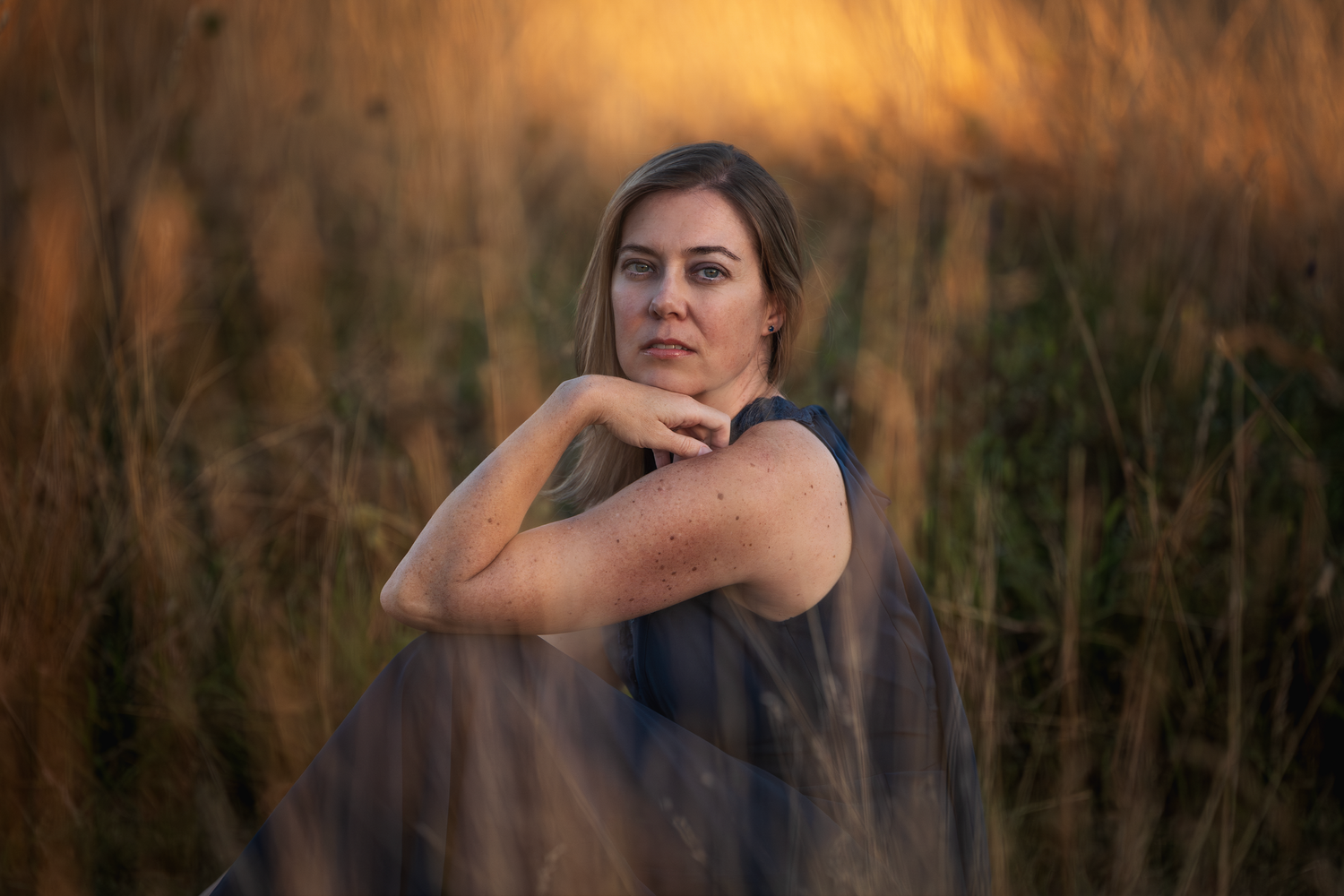 A woman in a navy dress poses while surrounded by tall grass with beautiful natural golden light of a sunset.