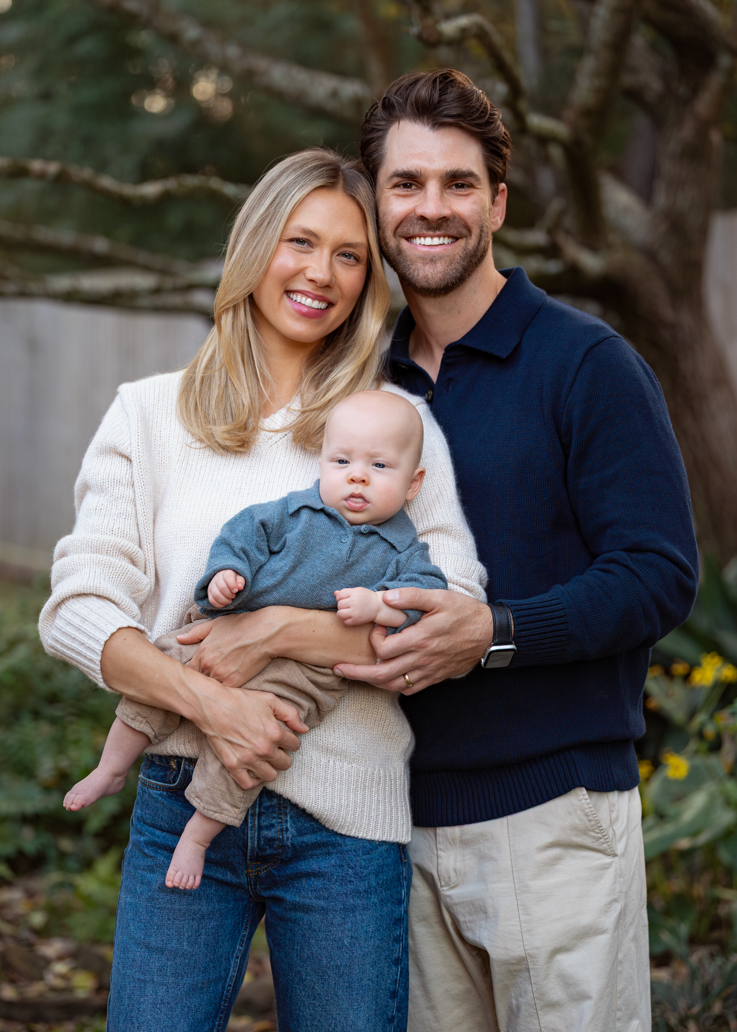 Smiling couple holding a baby infant outdoors in a garden setting, posing for a family portrait photo.