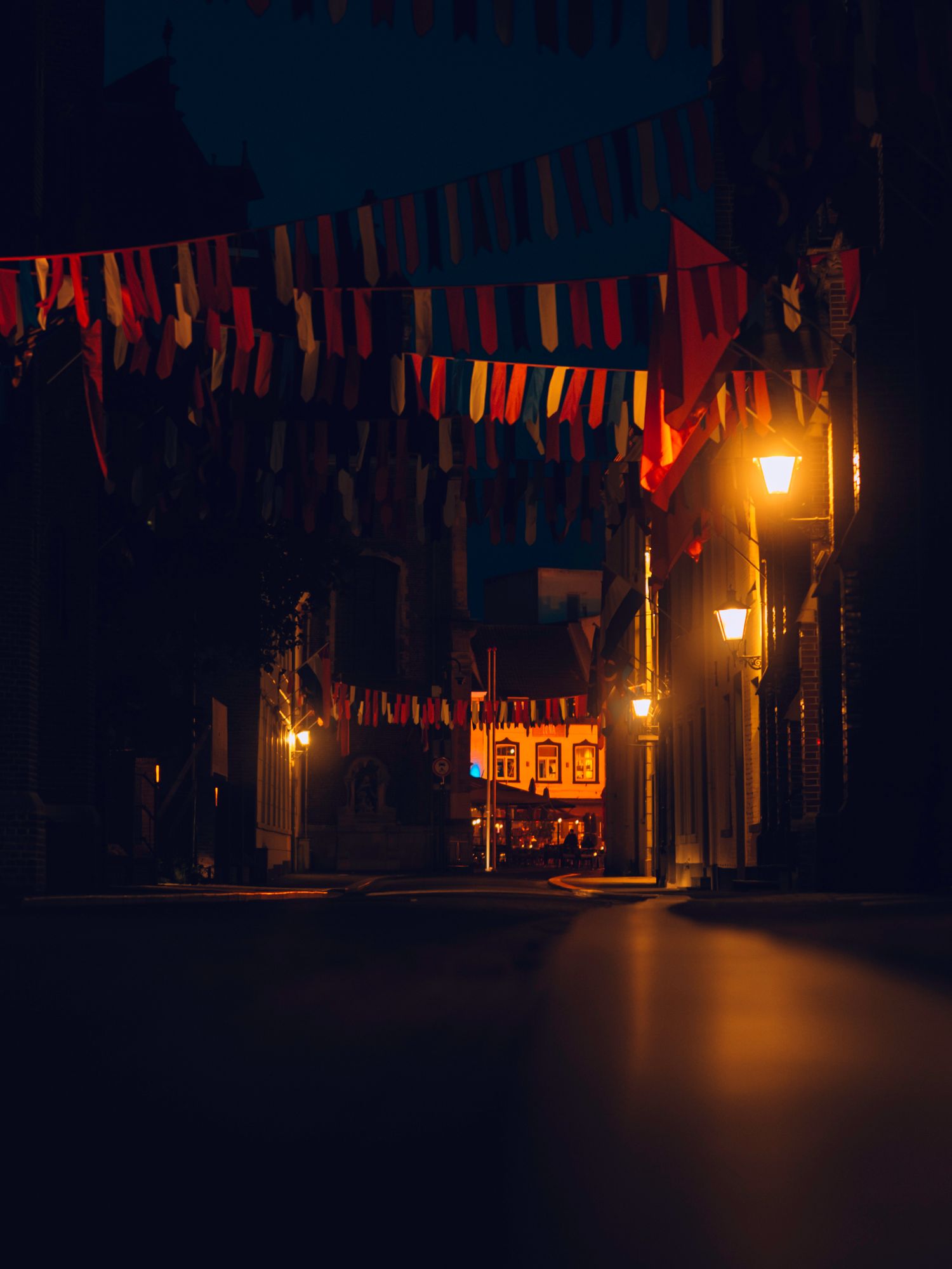 Colorful bunting flags hang over a dimly lit street at night with warm glowing lanterns creating an atmospheric scene.