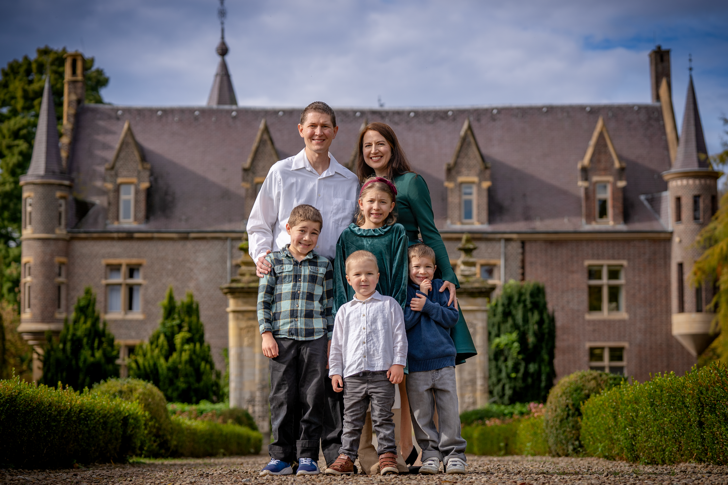 Family of six posing in front of a grand castle estate surrounded by lush greenery on a cloudy day.