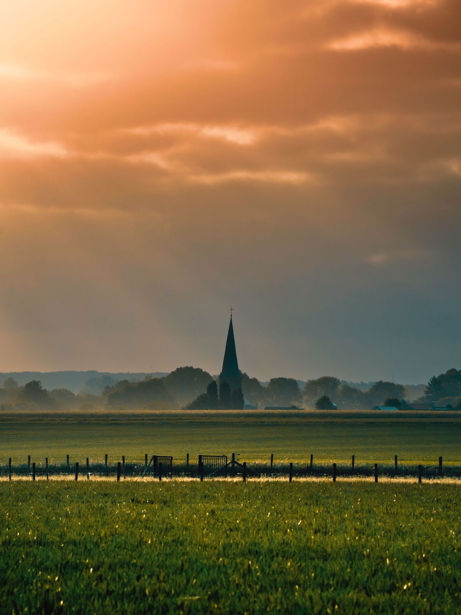 A panoramic view of a green meadow with a church spire silhouetted against a dramatic orange and teal sunset sky.