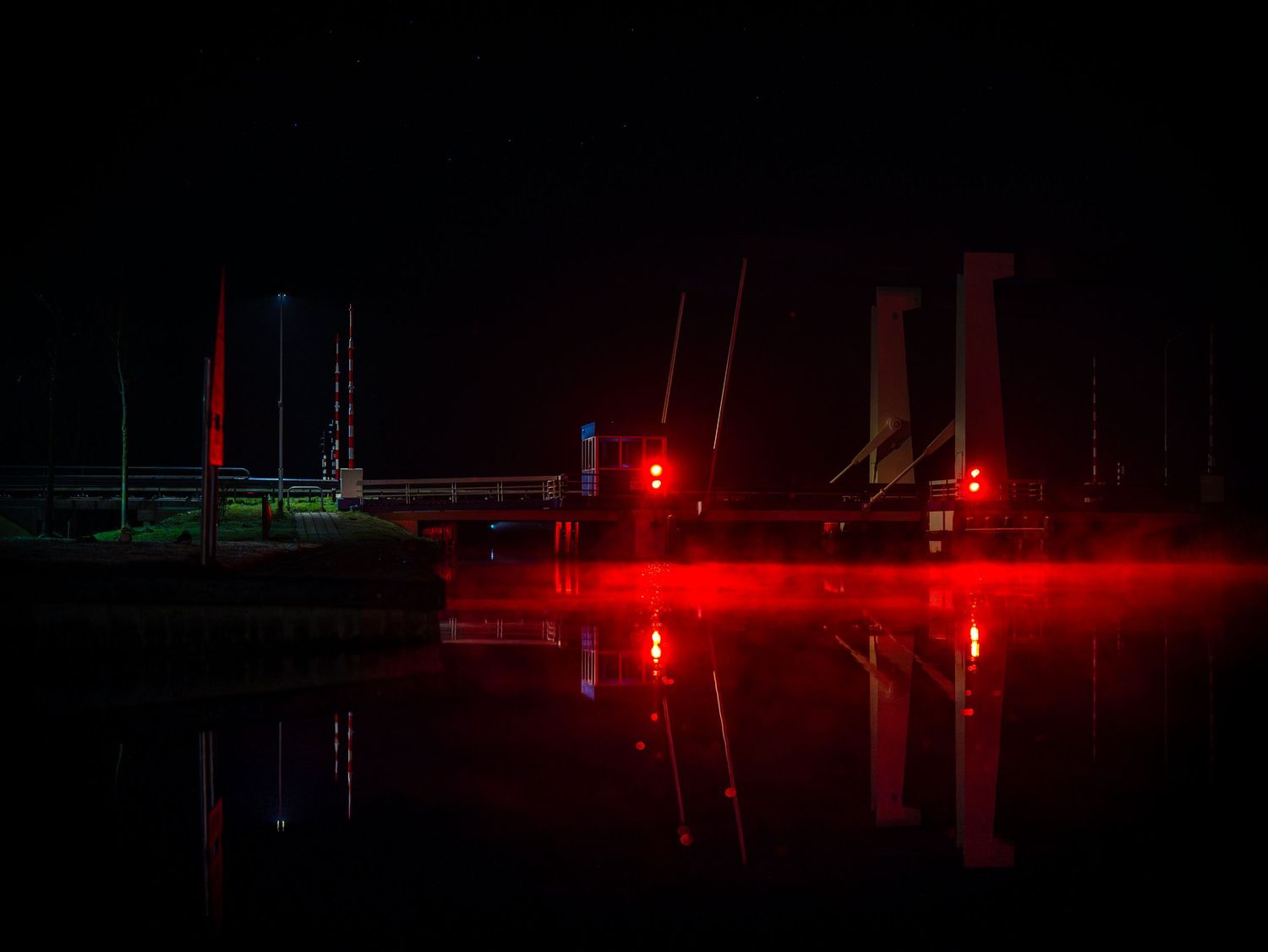 Bridge at night with red warning lights reflecting on water surface creating an eerie atmosphere.