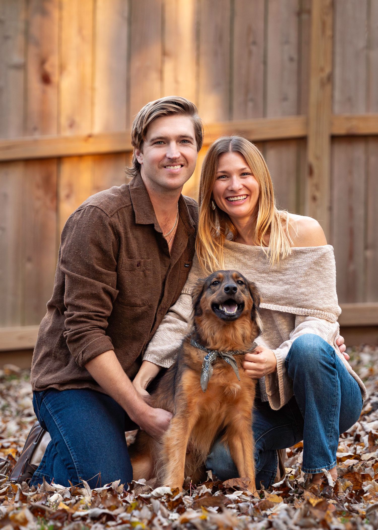 Smiling couple poses with happy brown dog on autumn leaves in front of wooden fence.