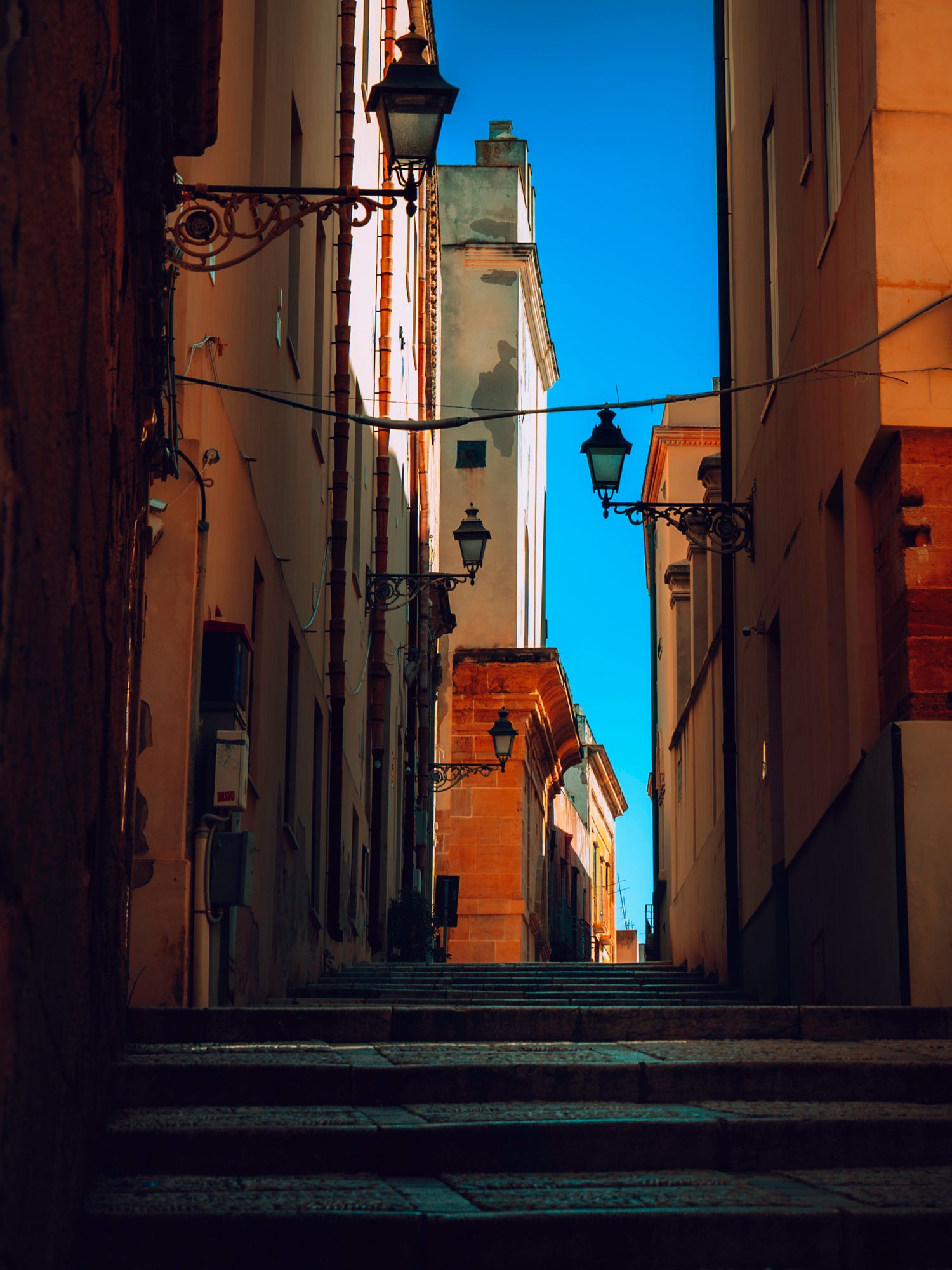 Narrow cobblestone alley with vintage street lamps and orange-tinted walls in an old Italian town during late afternoon.