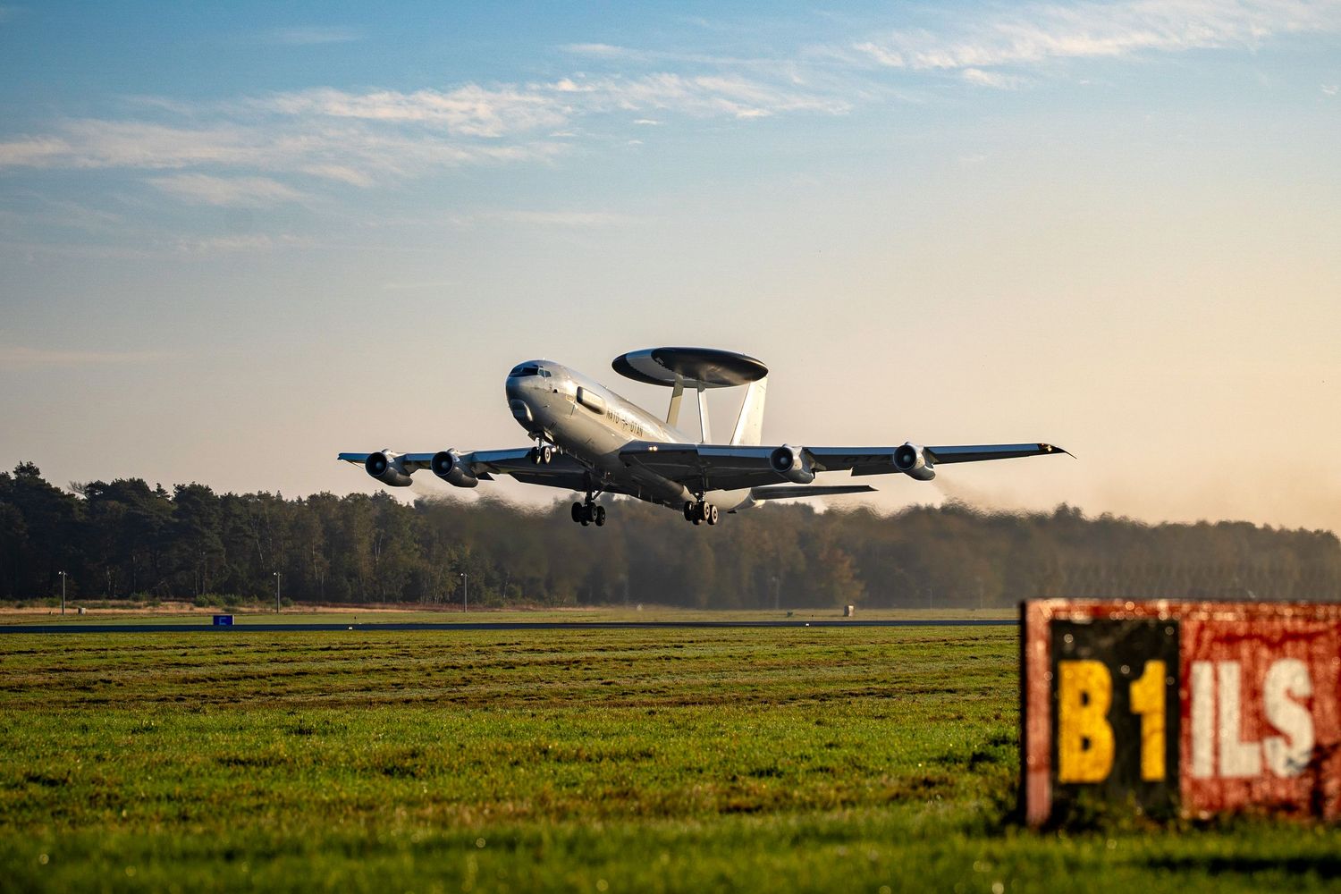 Military surveillance aircraft takes off from grassy airfield at sunset against cloudy sky.