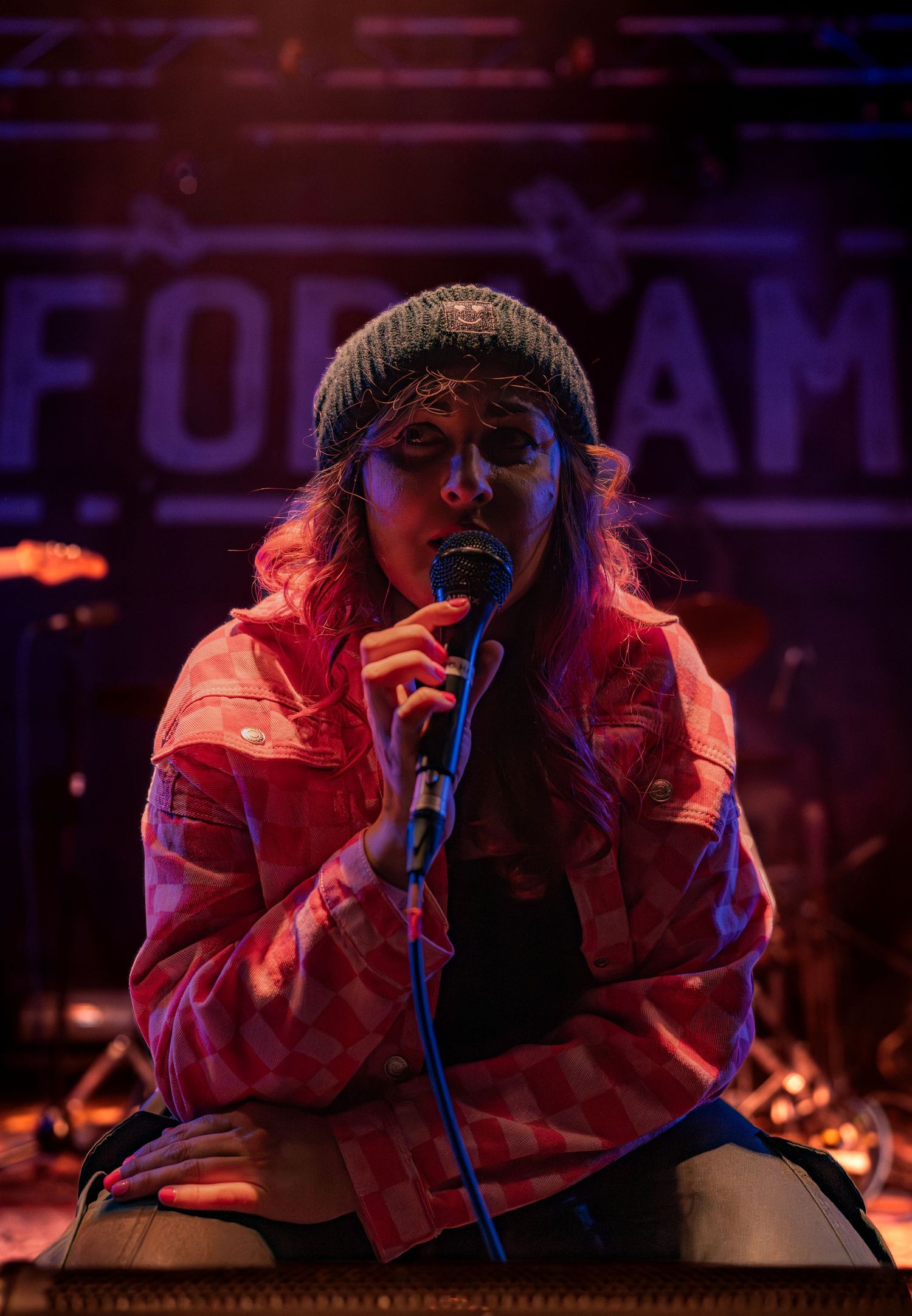 A performer in a pink jacket sings passionately into a microphone on a dimly lit stage during a concert performance.