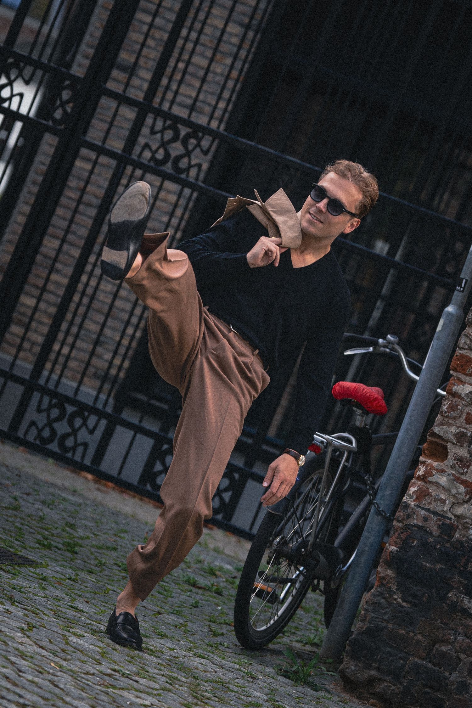 A man kicks his leg up on a cobblestone street near a bicycle and iron gate.