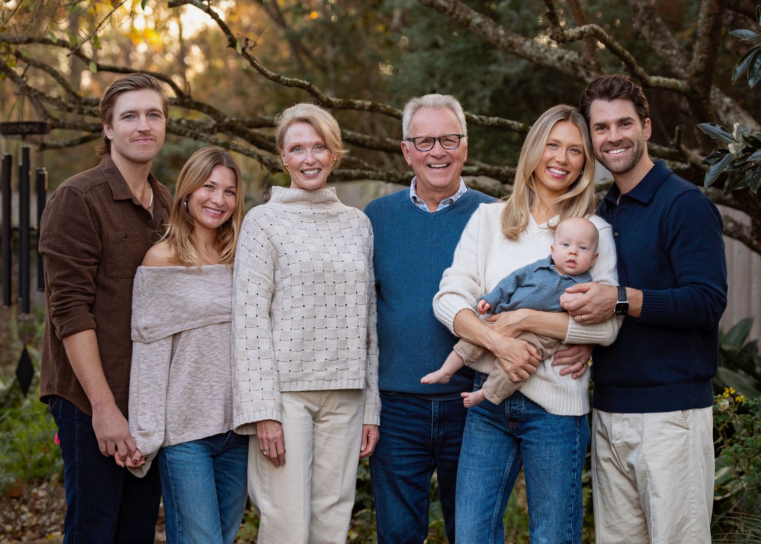 A joyful multigenerational family of six poses outdoors, including a baby, smiling warmly in a lush garden setting.
