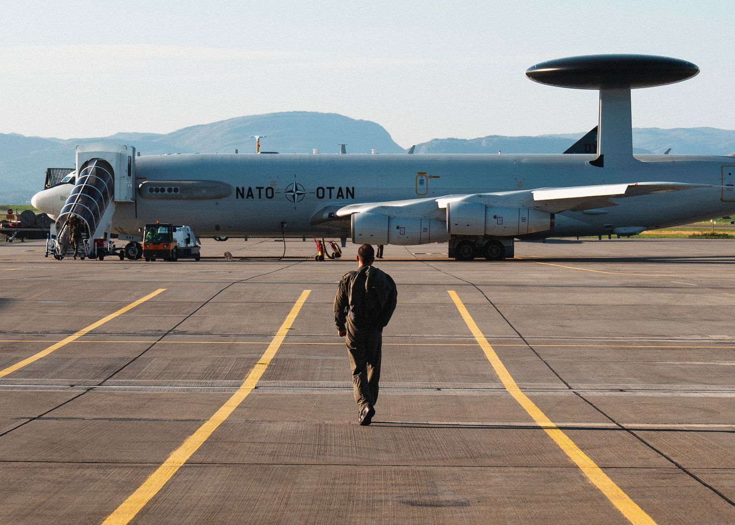 Military personnel walk across an airfield with a NATO AWACS E-3A aircraft parked in the background.
