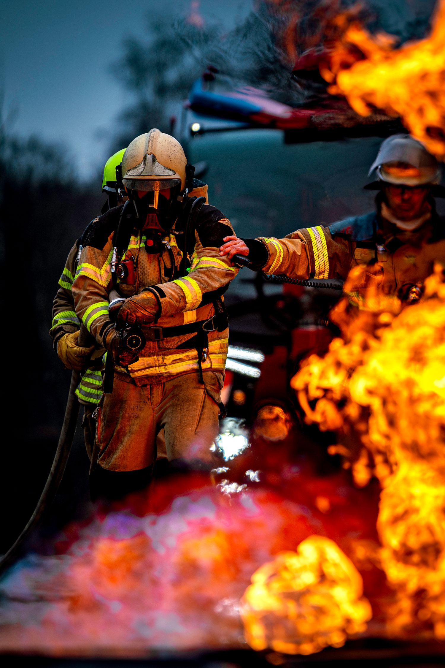 Firefighters in protective gear battle intense orange flames against a dark background during an emergency response simulation.