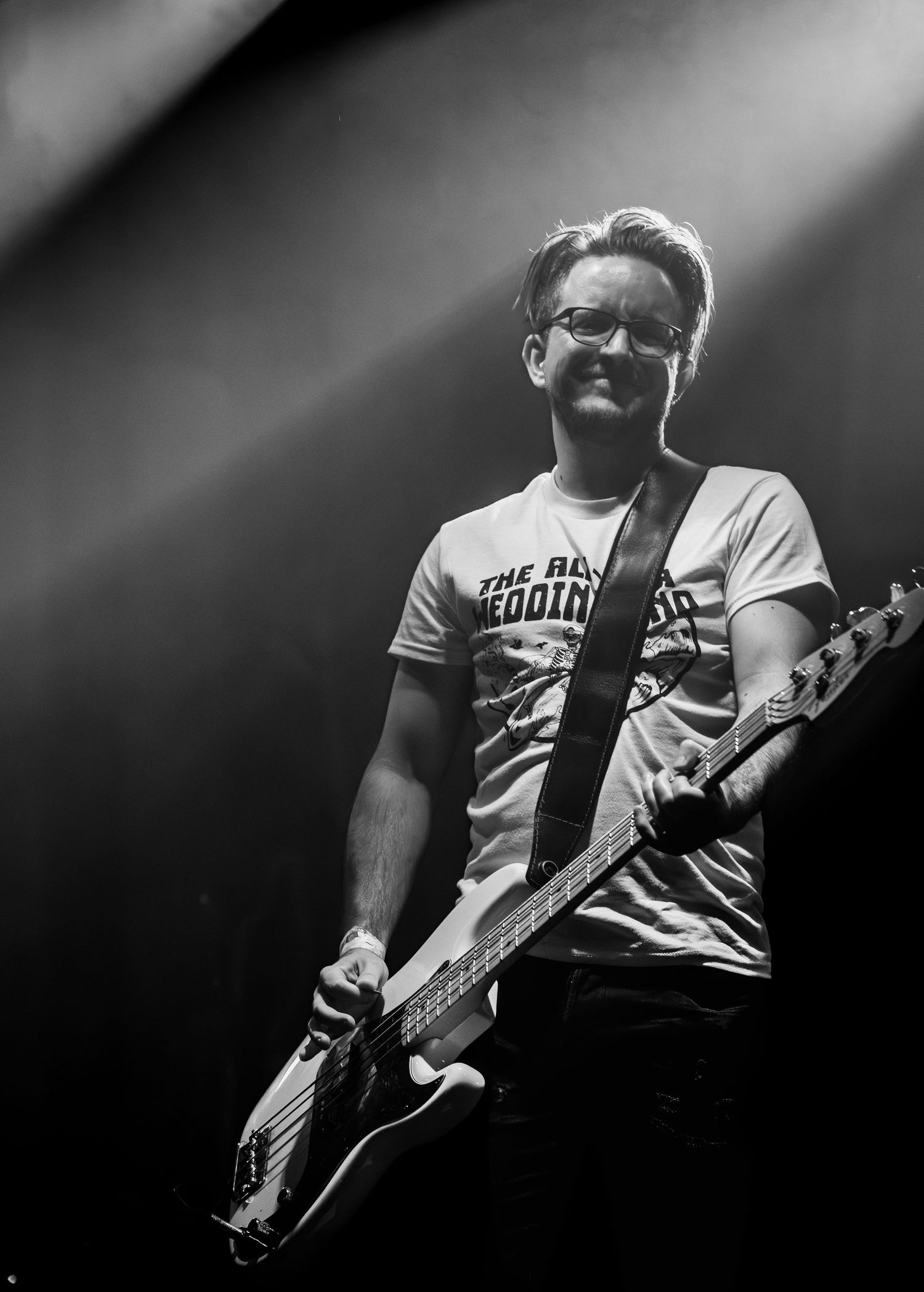 Black and white concert photo showing a musician playing bass guitar on stage in dramatic lighting.