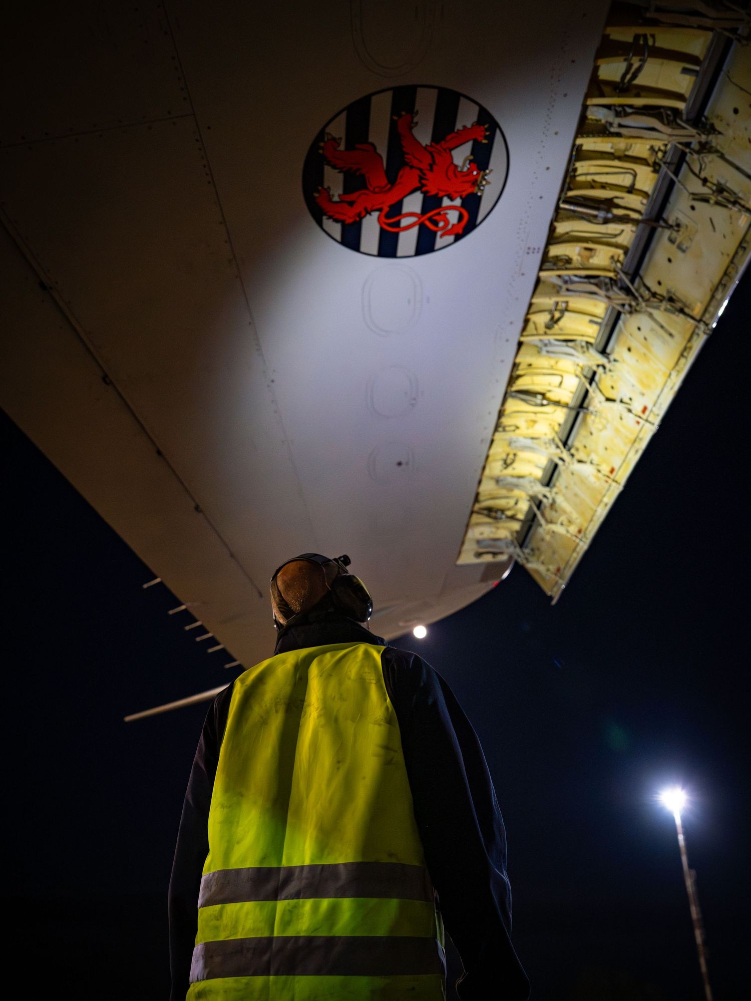 Worker in yellow safety vest inspects a NATO AWACS E-3A aircraft at night with a flashlight.