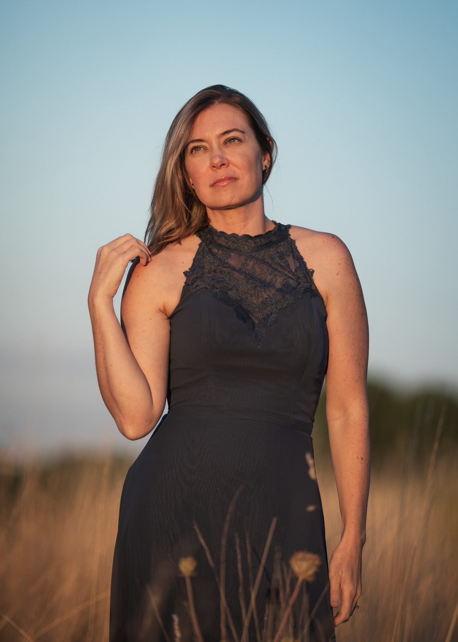 Woman in black lace halter dress standing in golden field at sunset with blue sky background.