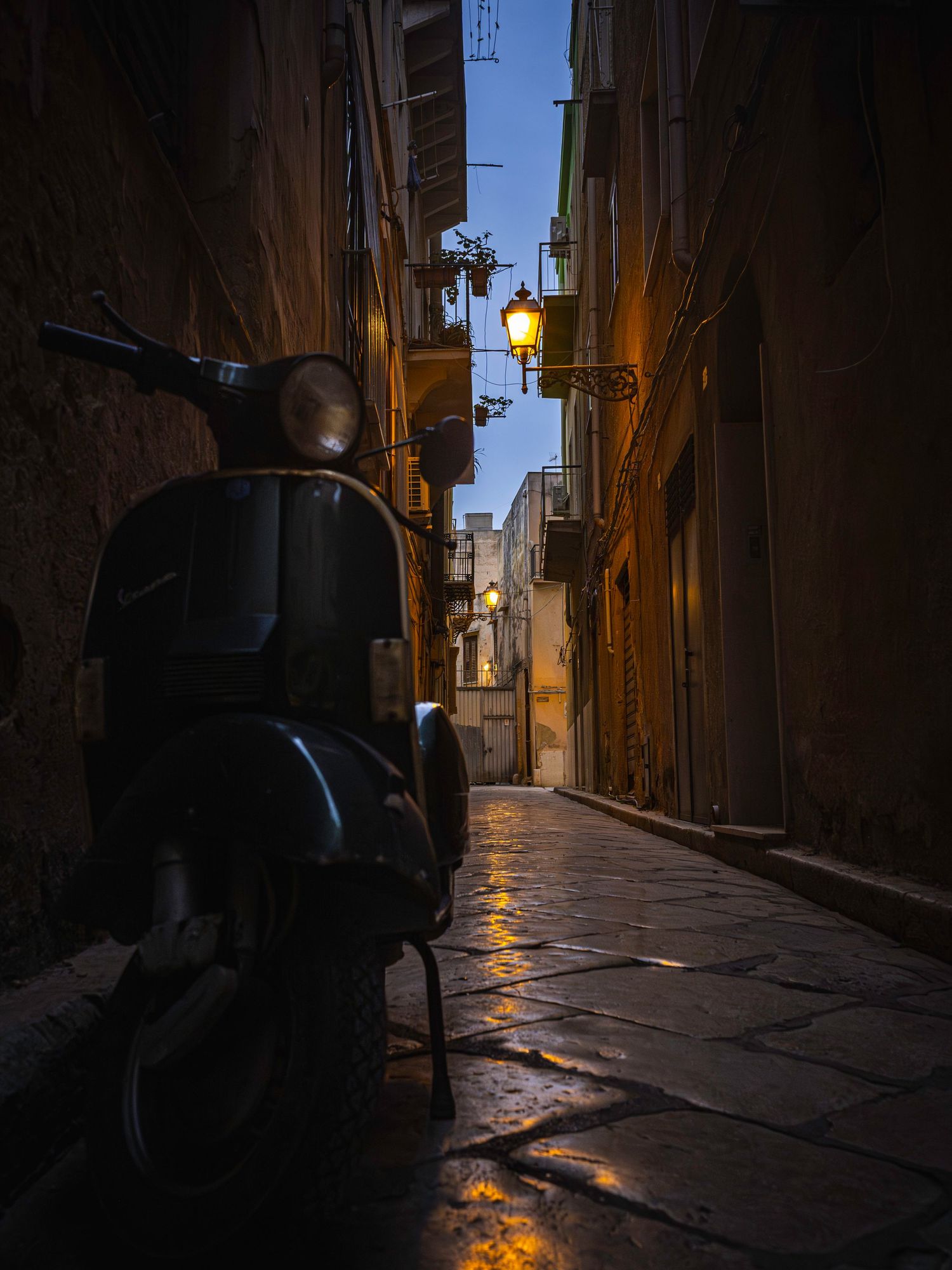 A vintage Vespa scooter parked in a narrow cobblestone alley of an Italian city at dusk with glowing street lamps.