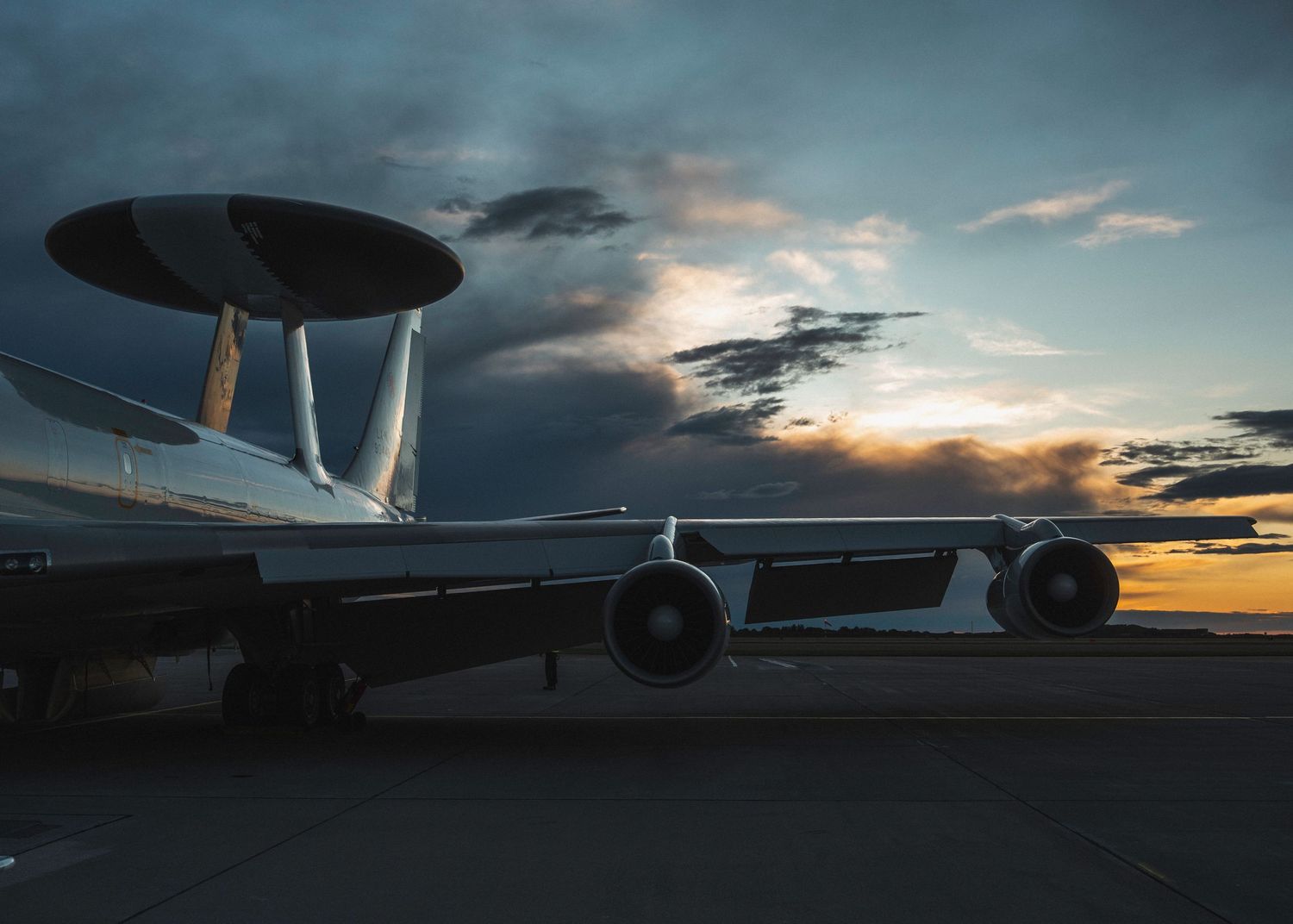 Military aircraft silhouetted against dramatic sunset sky with dark clouds and orange horizon glow.