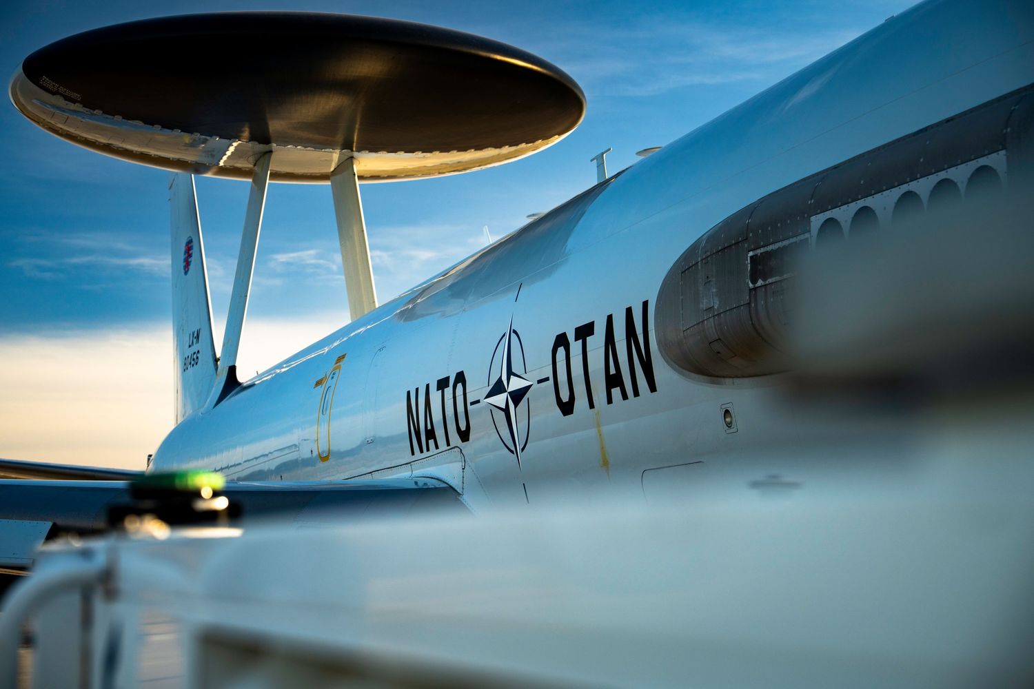 A close-up view of a NATO AWACS E-3A aircraft radome and fuselage against a bright blue sky.