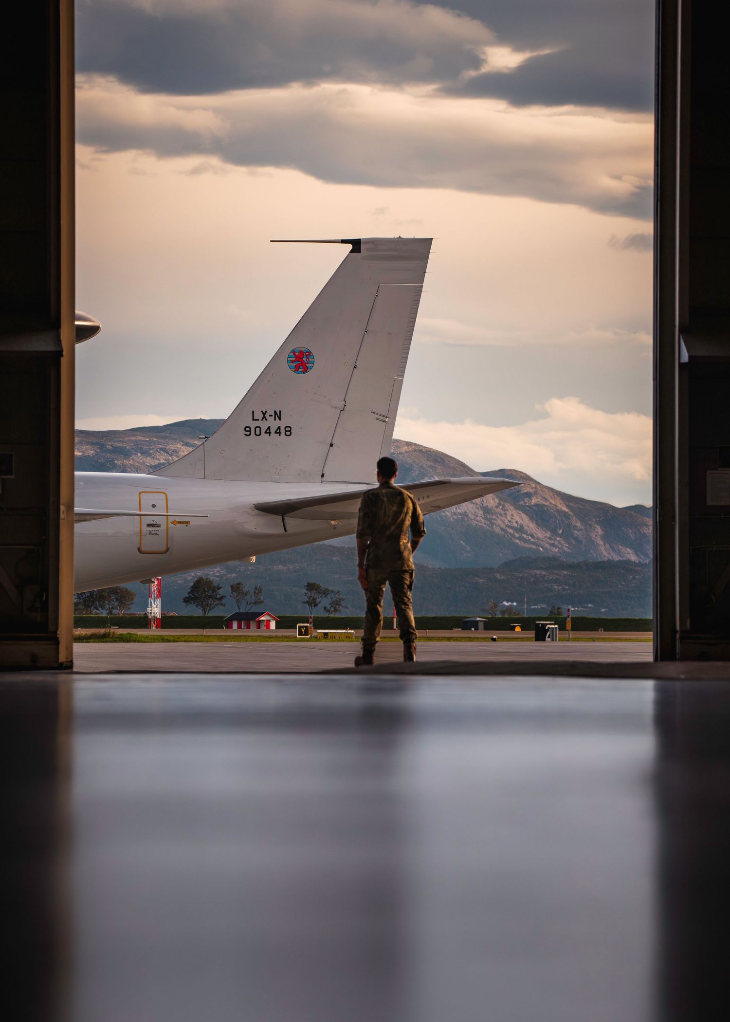 A silhouetted figure stands near a parked aircraft on an airfield at sunset with mountains in the background.