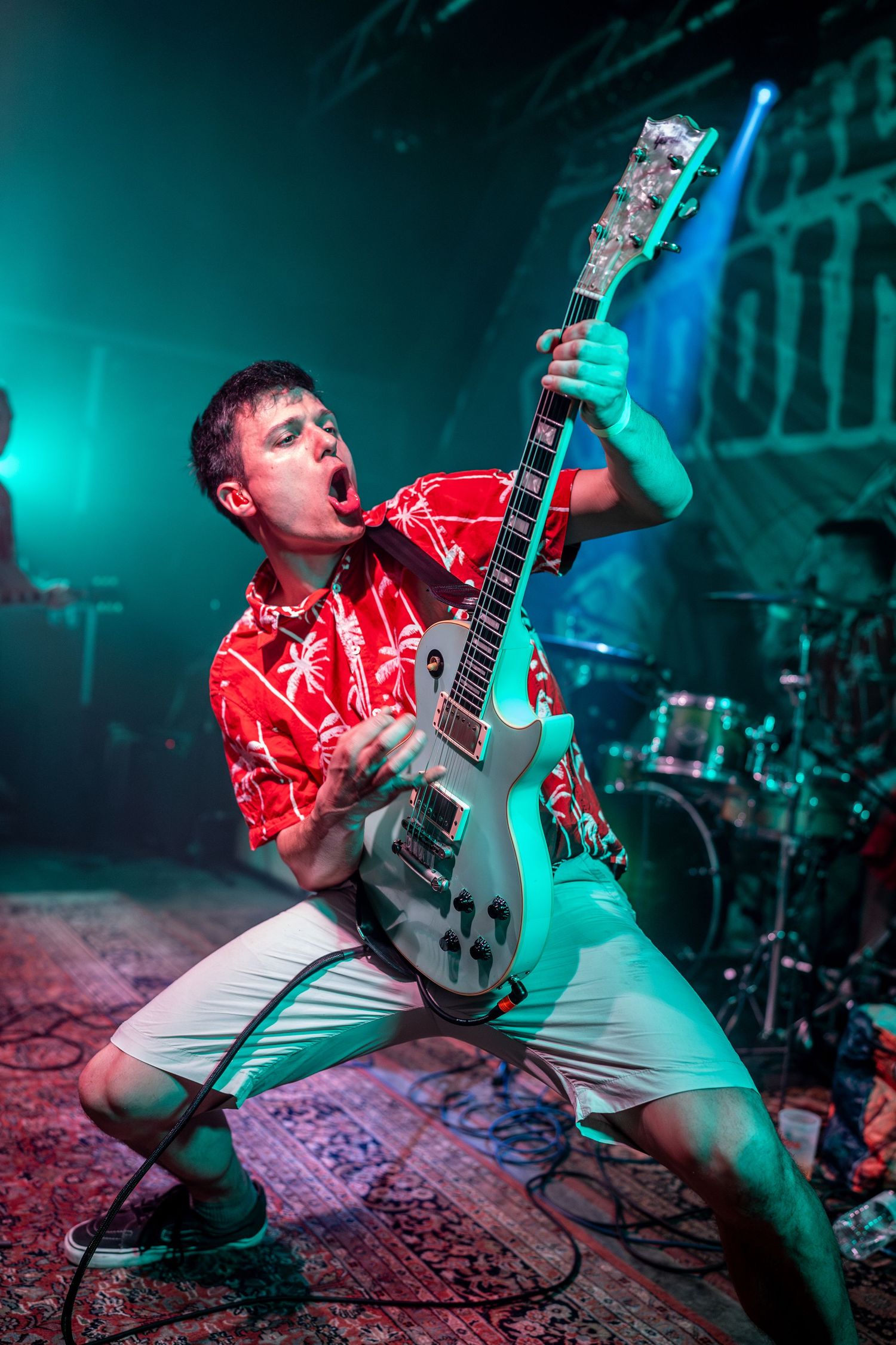 Energetic guitarist in red Hawaiian shirt plays white electric guitar on stage under teal lighting at live concert.