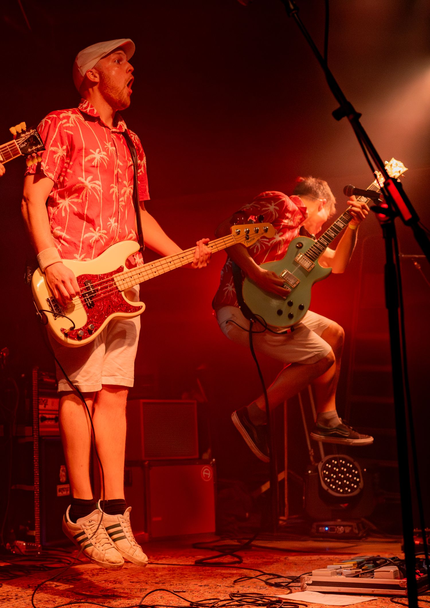 Musicians perform on stage in red lighting while wearing casual attire and playing the guitar and bass.