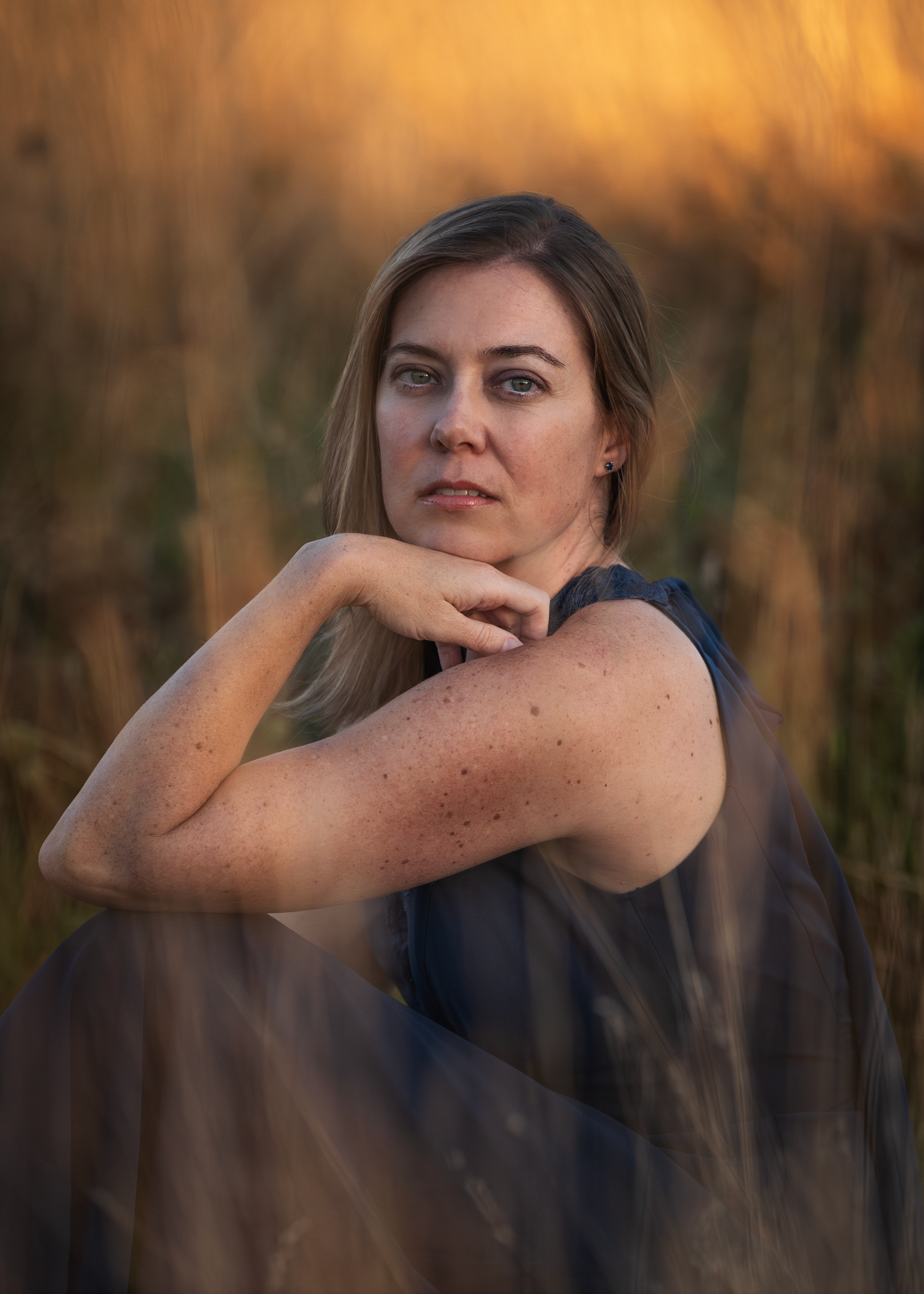 A woman in a blue dress sits thoughtfully in a field during golden hour lighting.