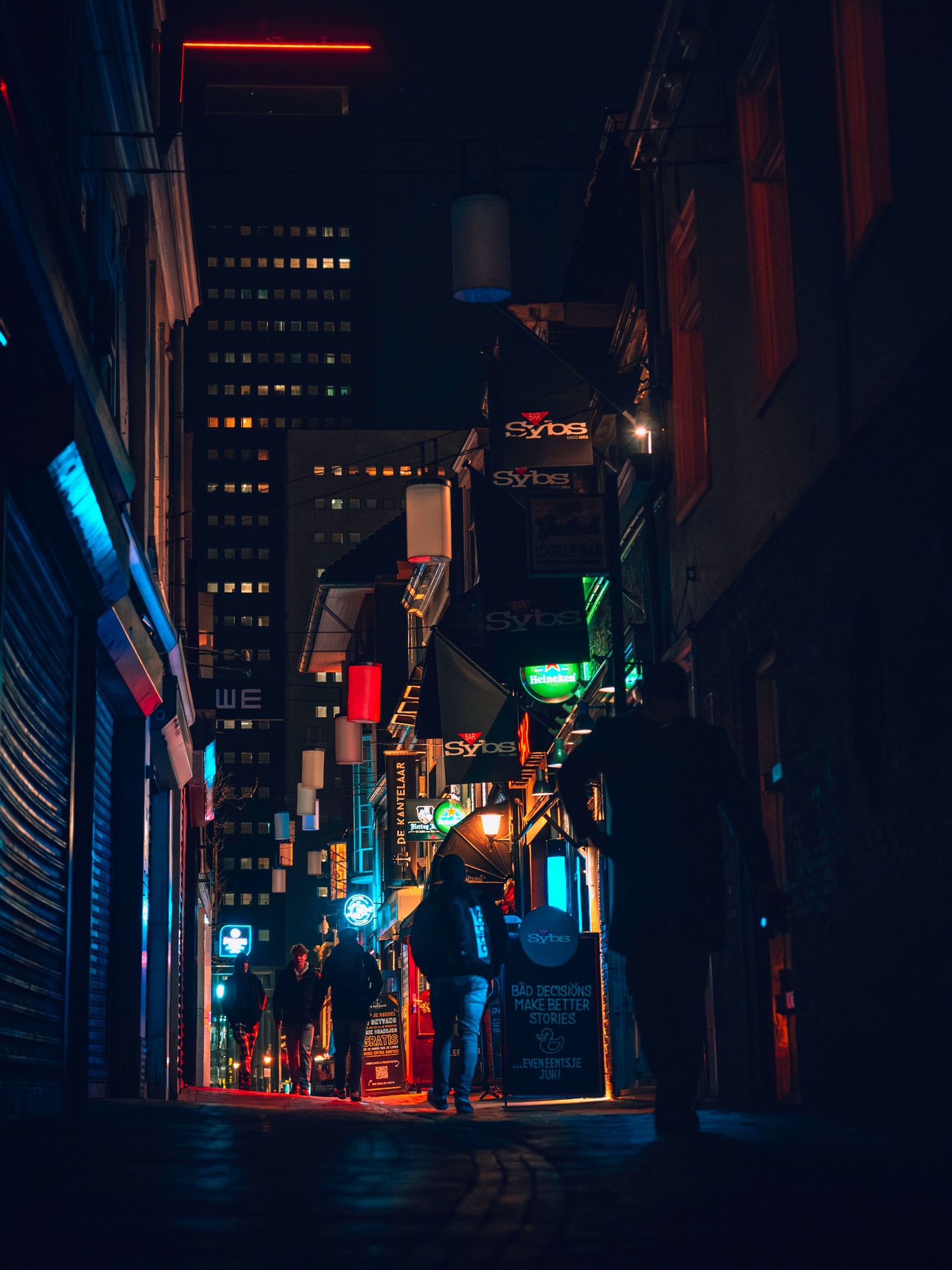 Moody urban alleyway at night illuminated by colorful neon lights casting red, blue and green glows on dark buildings.