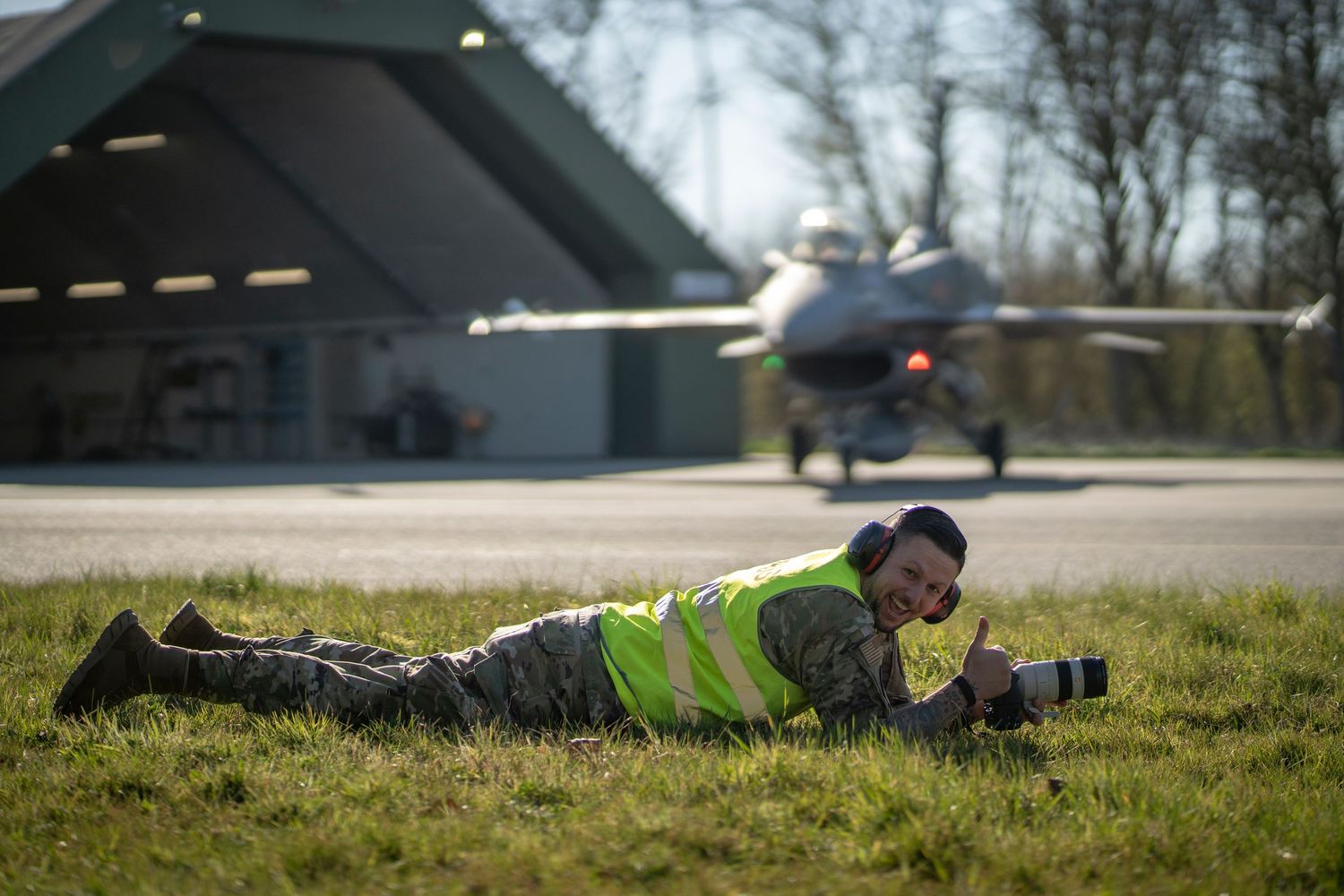 Aviation photographer in safety vest lies on grass to capture photos of aircraft at small airfield.