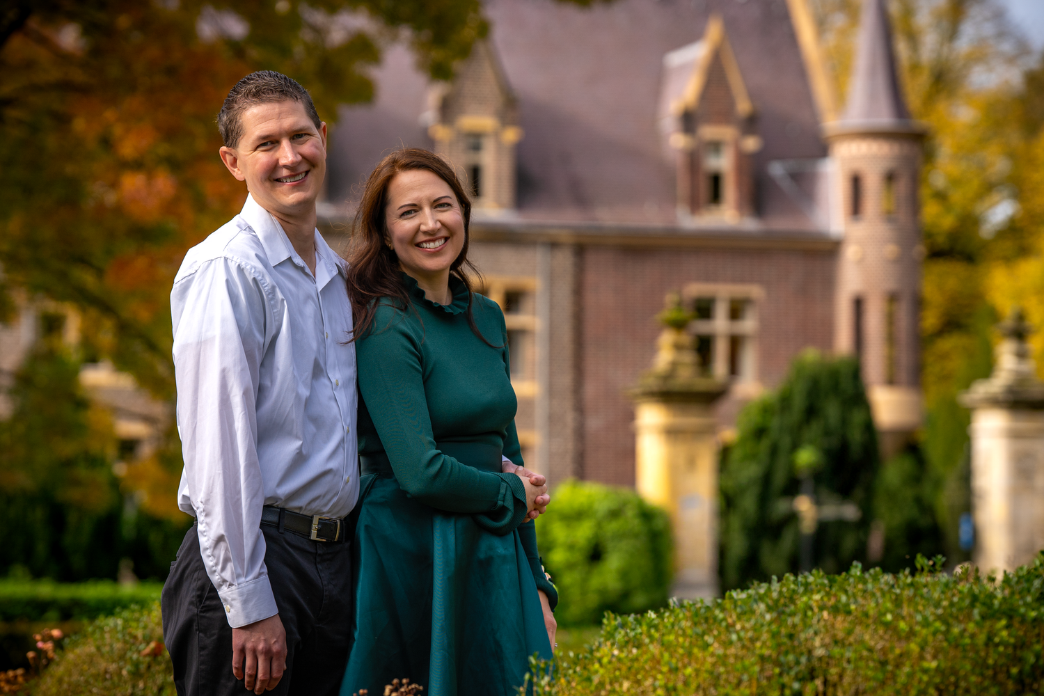 Couple smiling together in front of a historic stone castle surrounded by autumn foliage.