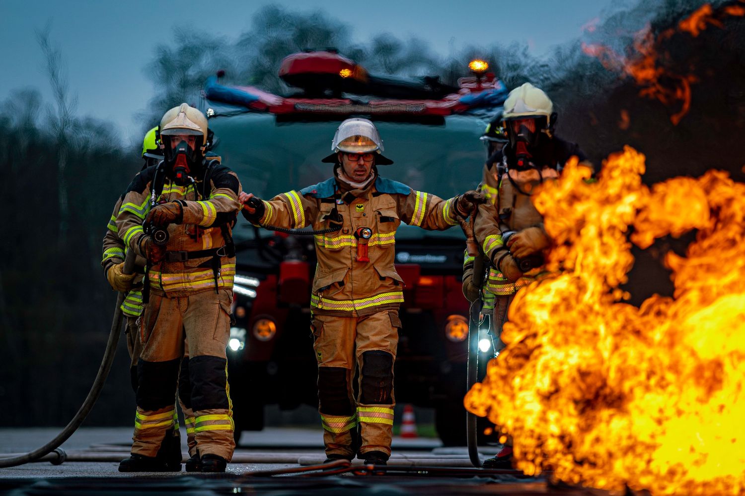 Firefighters in protective gear work together to control intense orange flames during a training exercise.
