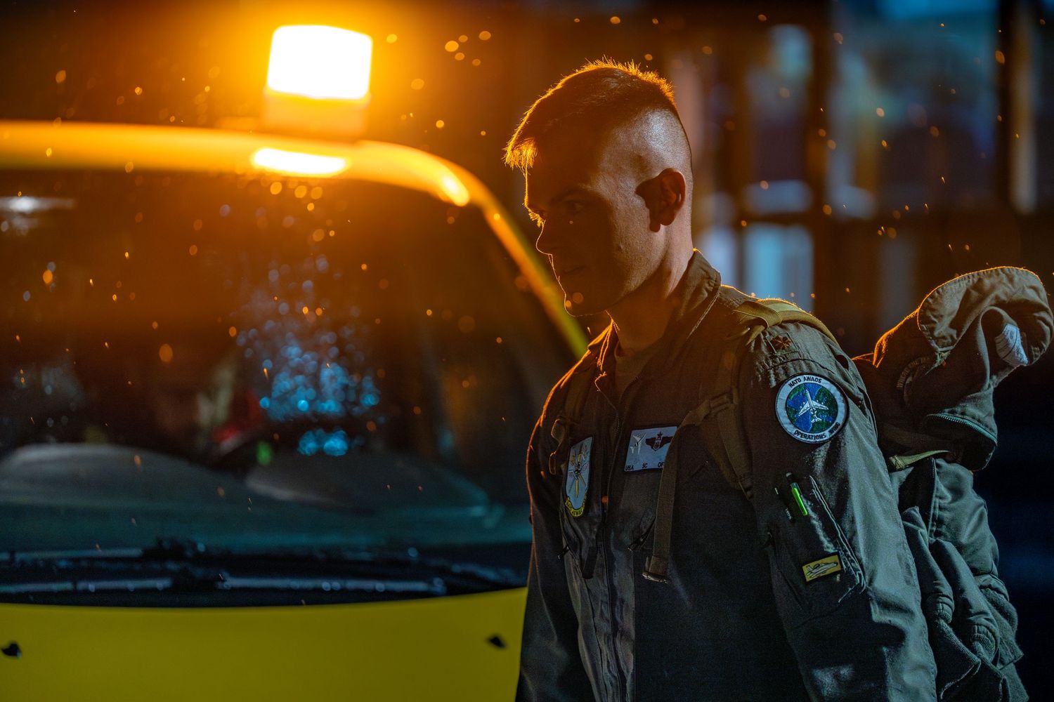 NATO AWACS crew member walking toward an AWACS aircraft for a mission, illuminated by vehicle warning lights at night.