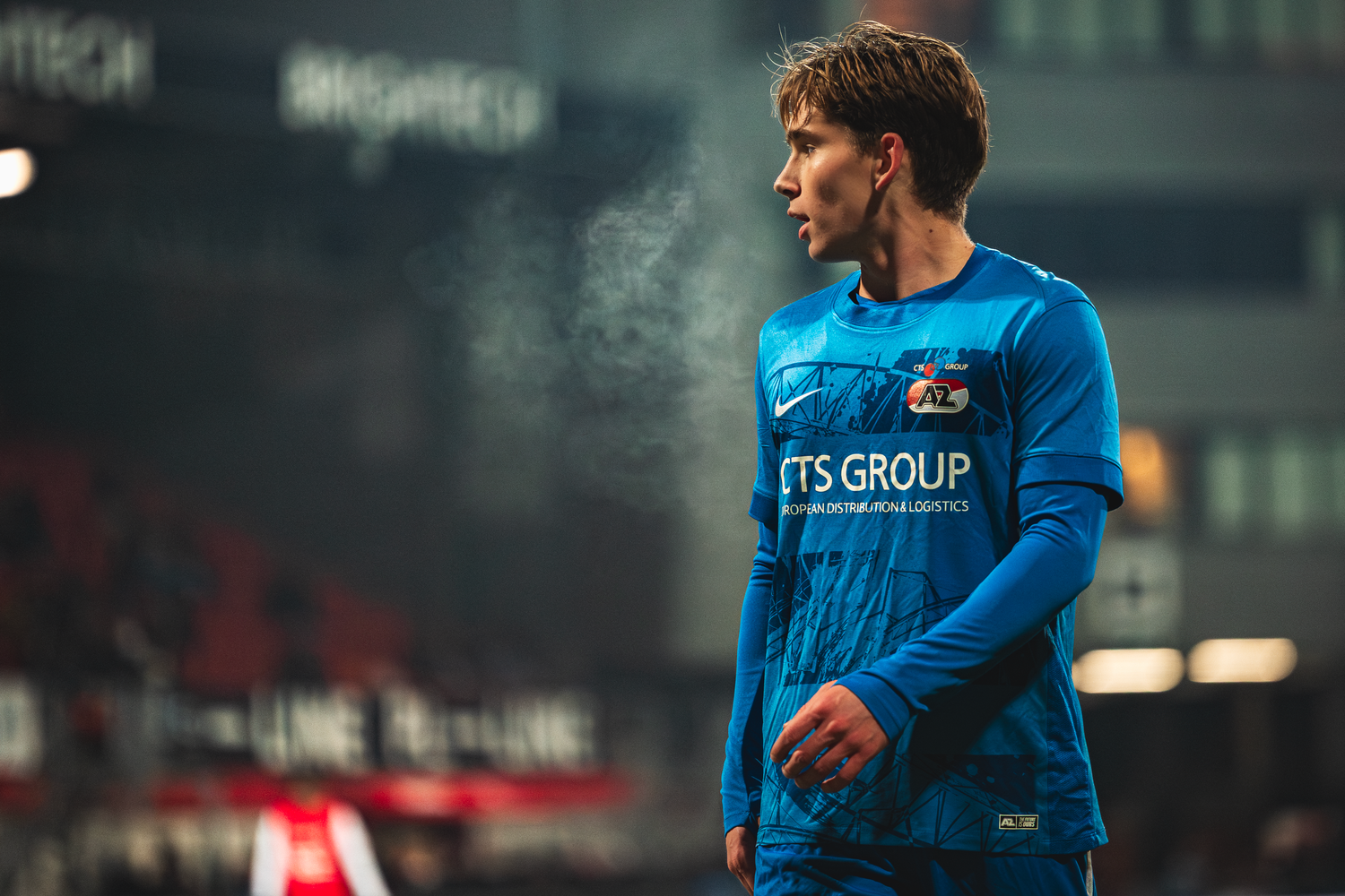 Soccer player wearing bright blue jersey with FTS Group sponsor logo stands on field during match.