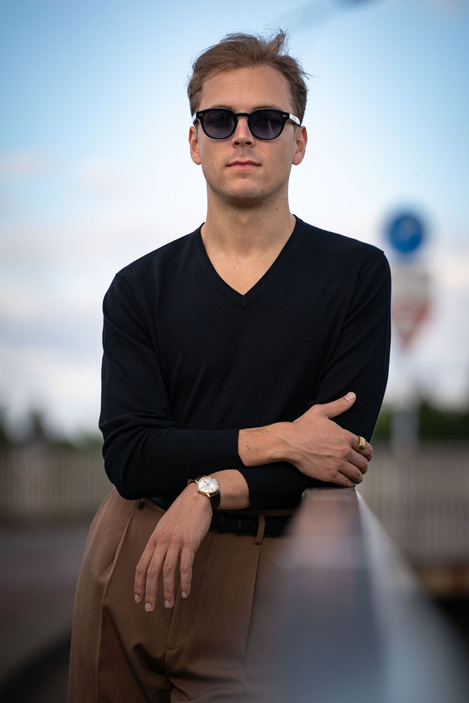 Portrait of a man in a black v-neck sweater and sunglasses with arms crossed, standing outdoors.