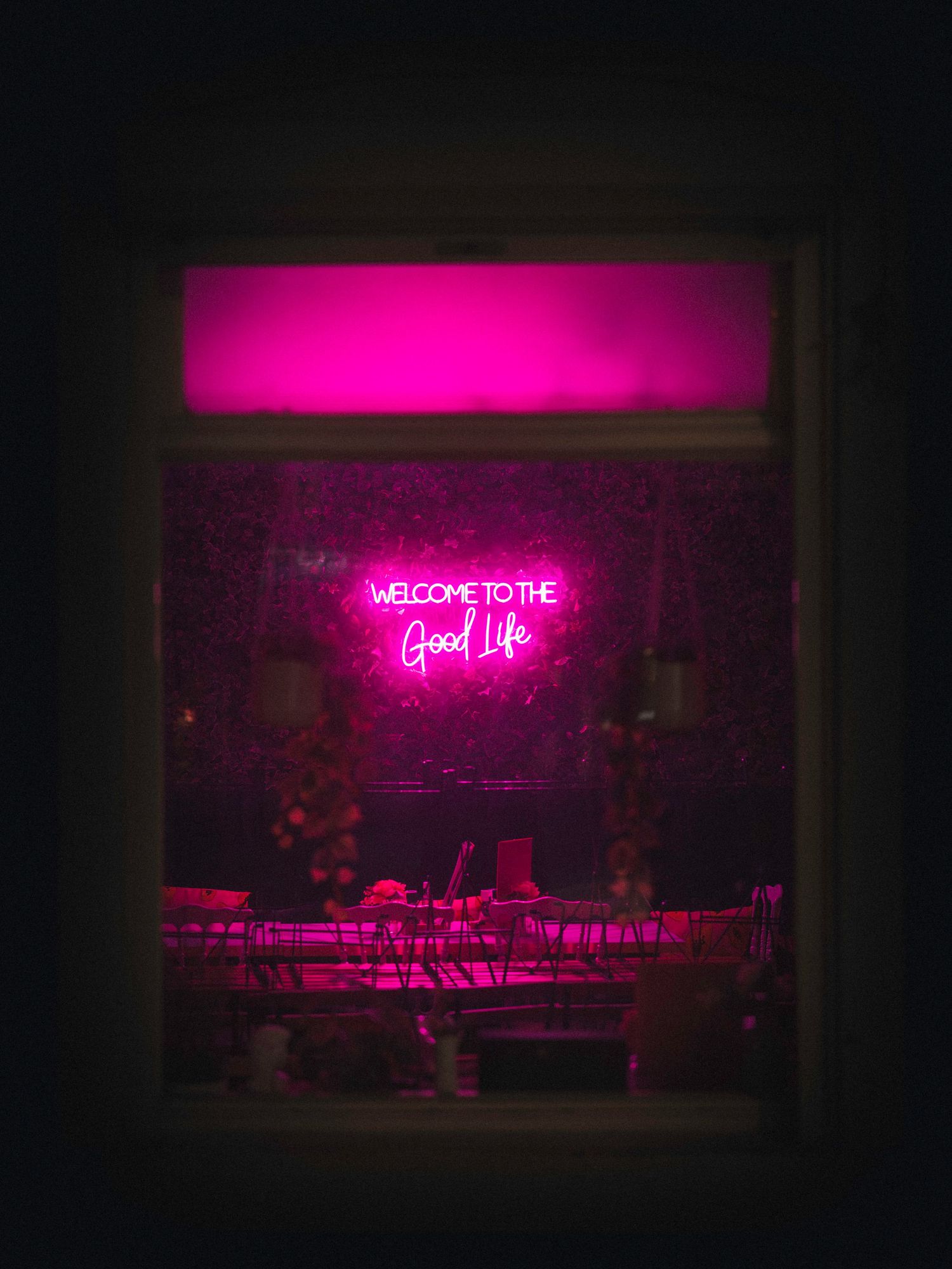 A series of pink neon signs in a dark room displaying text including 'Welcome to the Good Life' with bar seating.