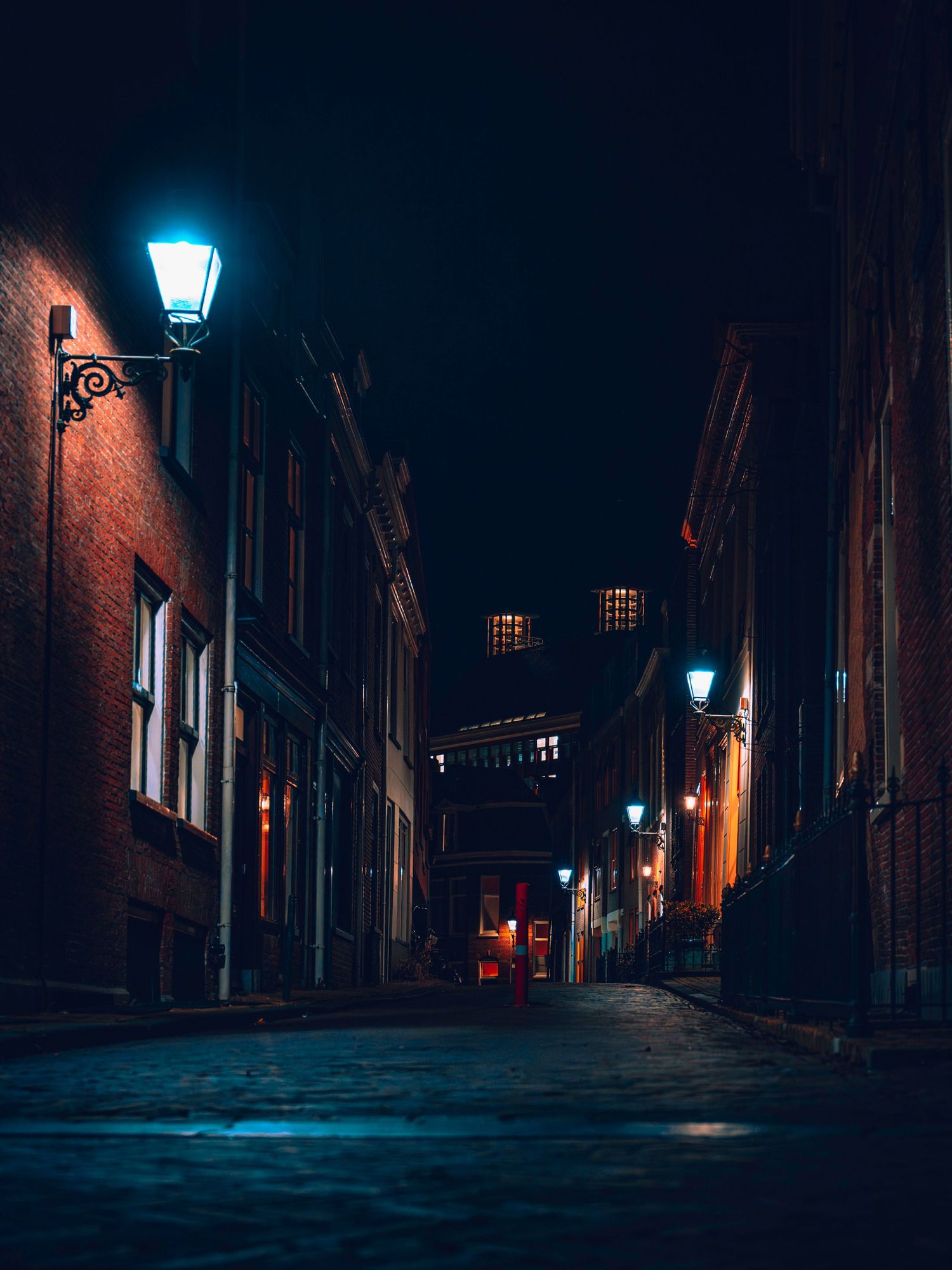 Moody nighttime street scene with glowing blue lamps illuminating narrow cobblestone alleyways in an old city quarter.