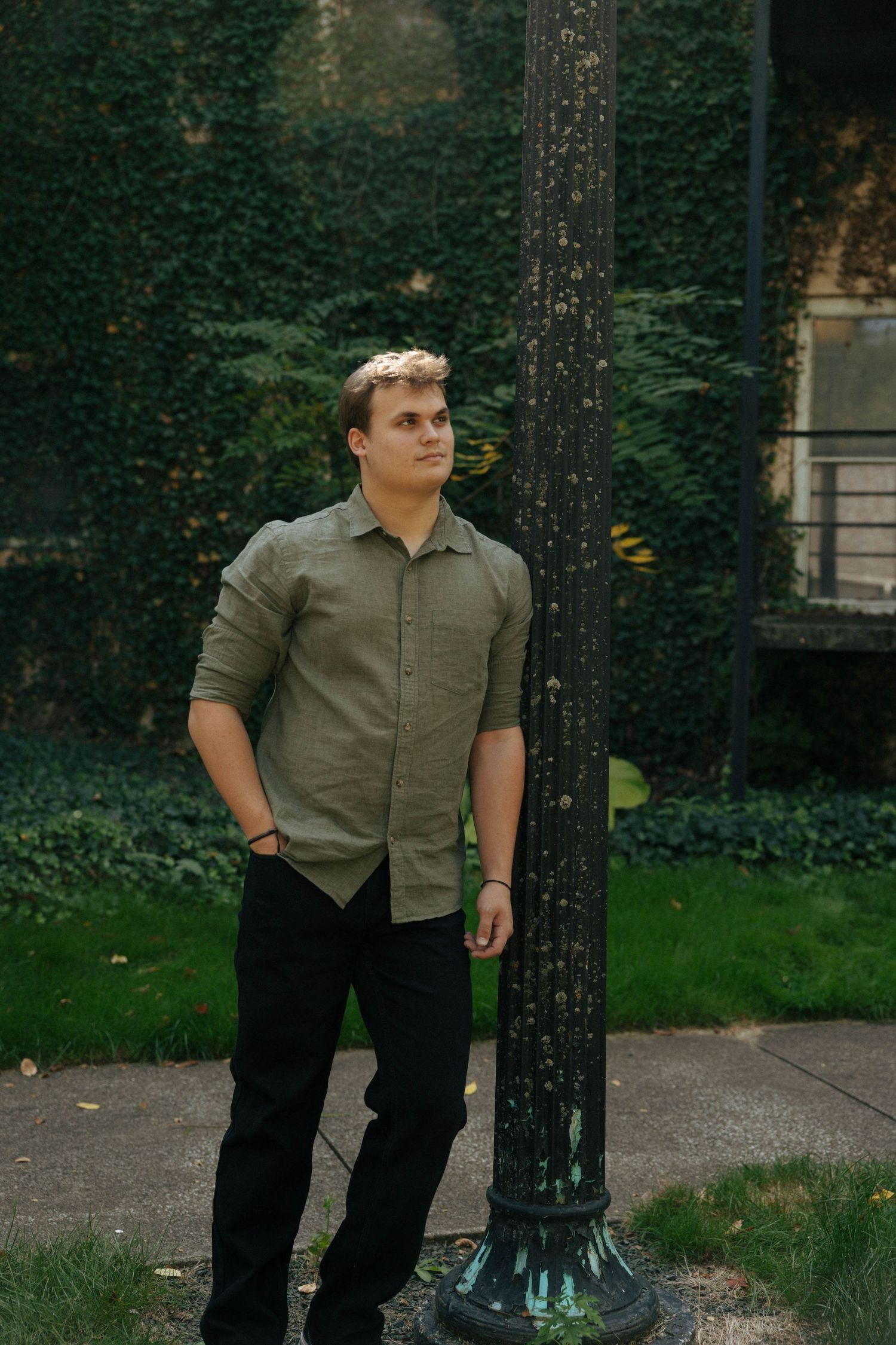 Person in olive shirt poses against dark tree backdrop in garden setting.