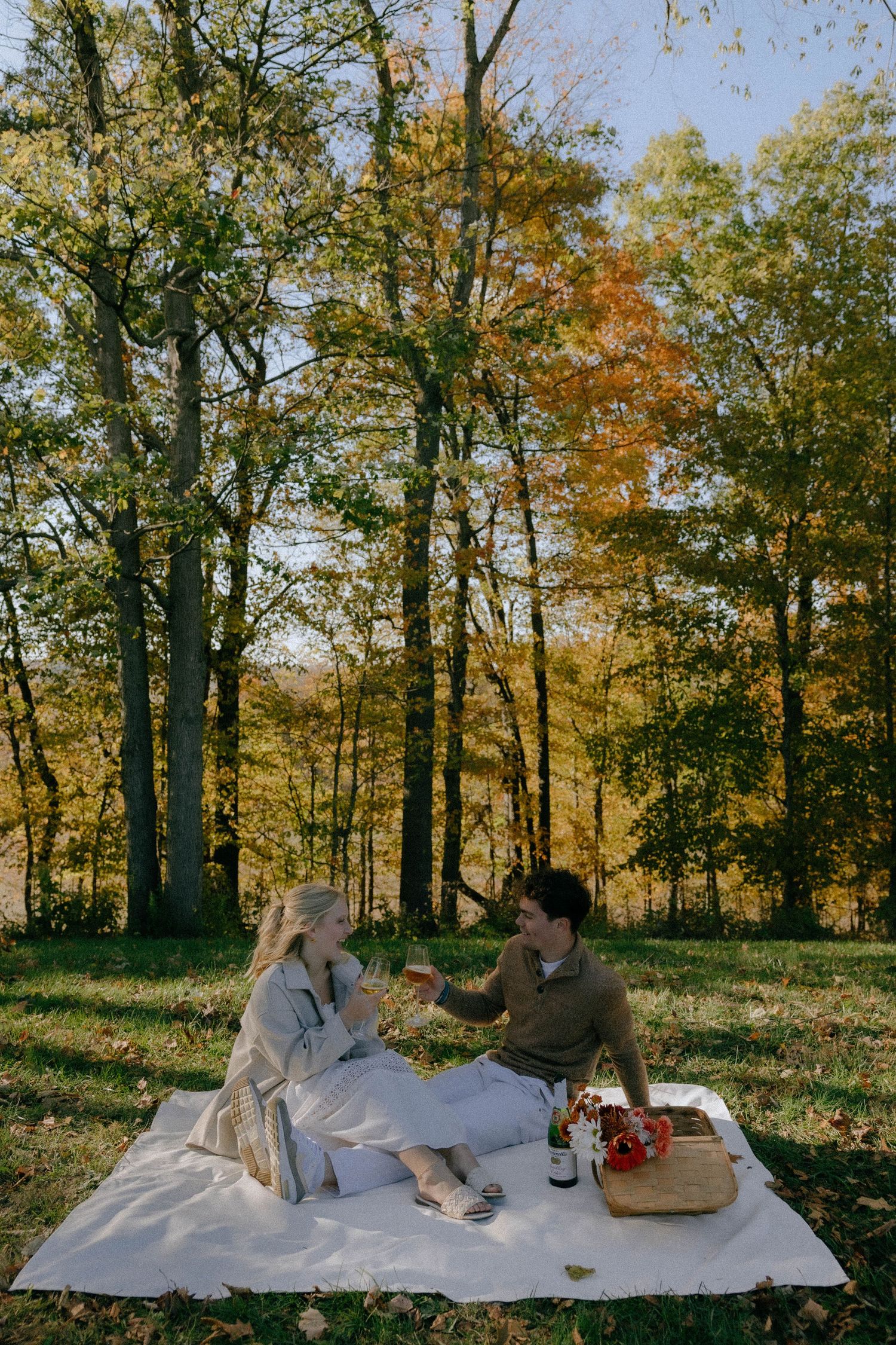 A couple enjoying wine and snacks on a white picnic blanket in a sunny autumn forest.