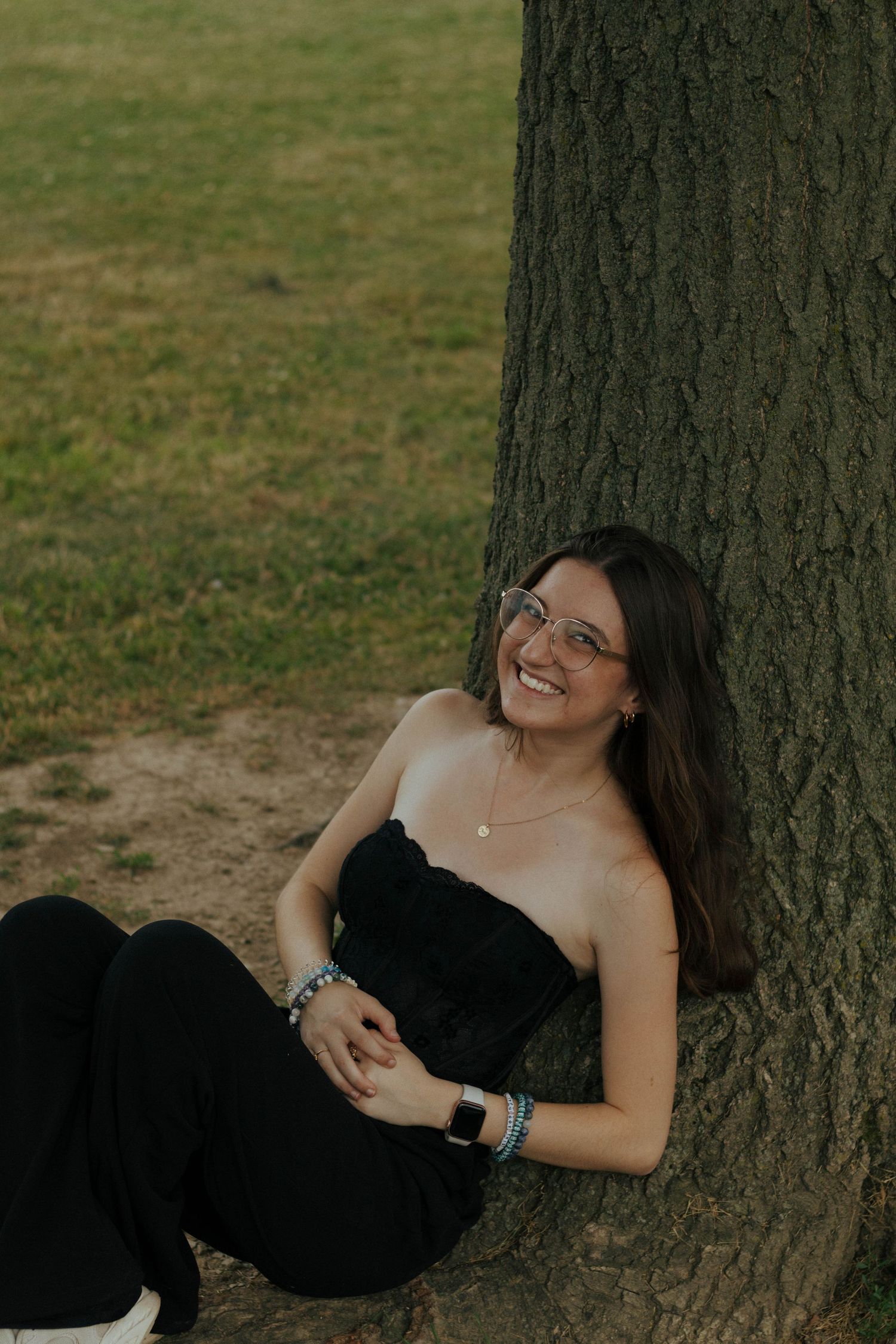 Woman in black dress reclining against a tree trunk in casual outdoor portrait.