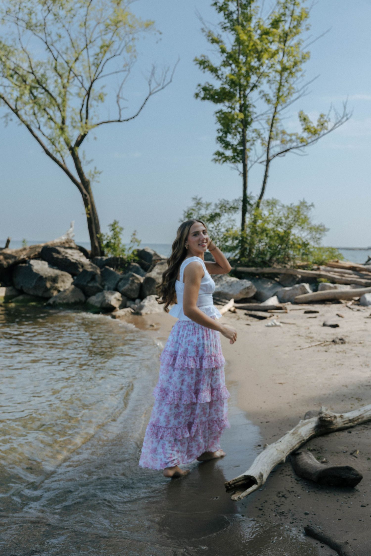Person in flowing dress poses on rocky beach shore near water.
