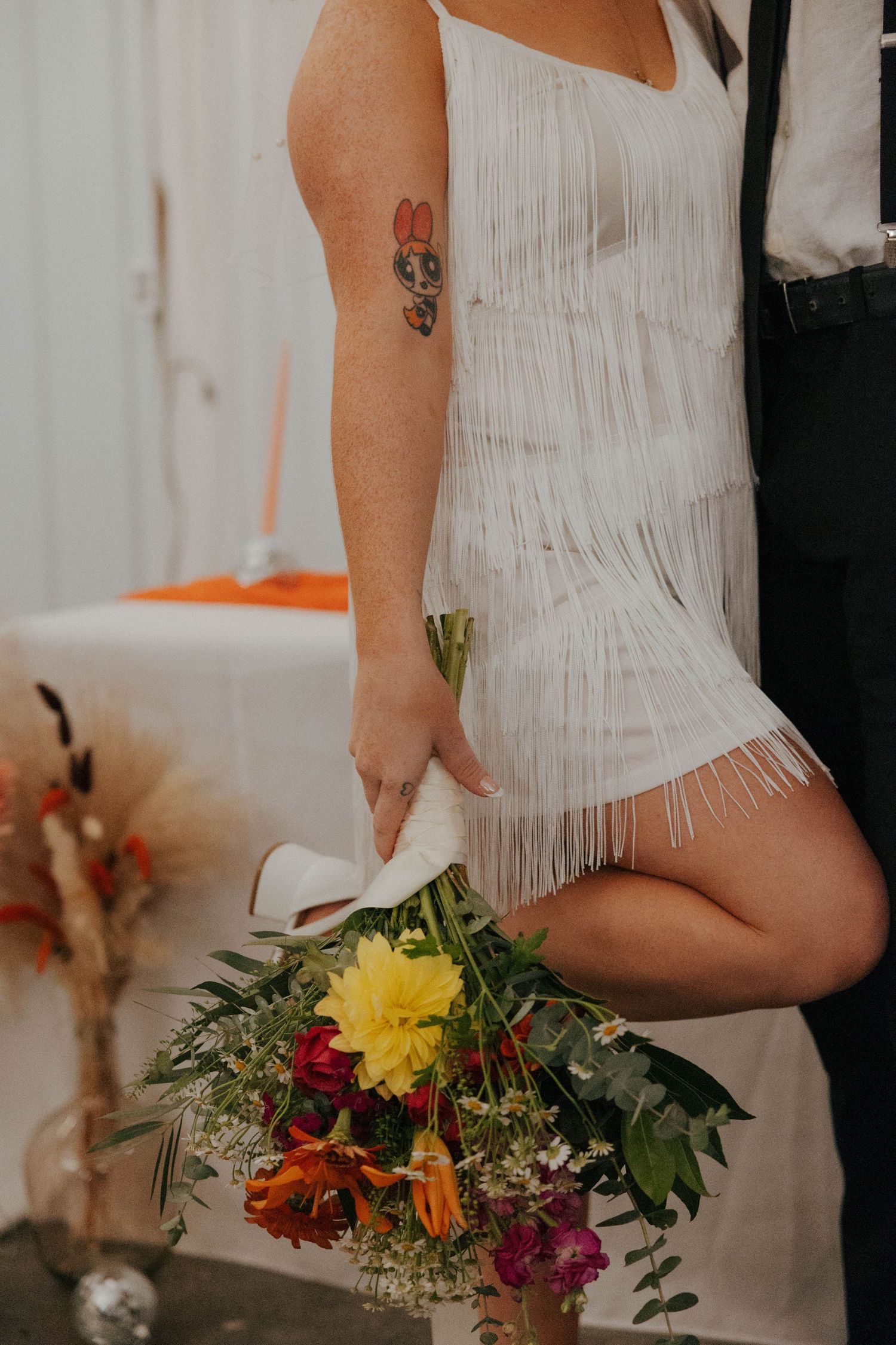 A white fringe dress detail with colorful wildflower bouquet against a neutral background.