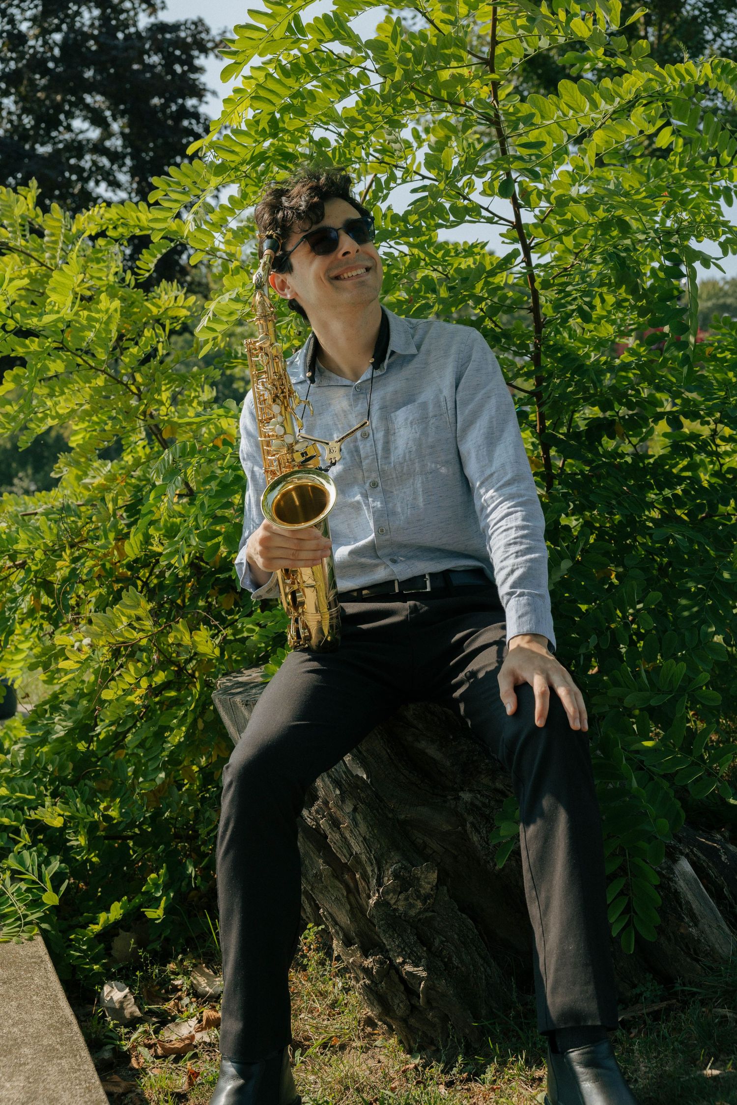 Saxophonist performing outdoors against a backdrop of lush green trees.