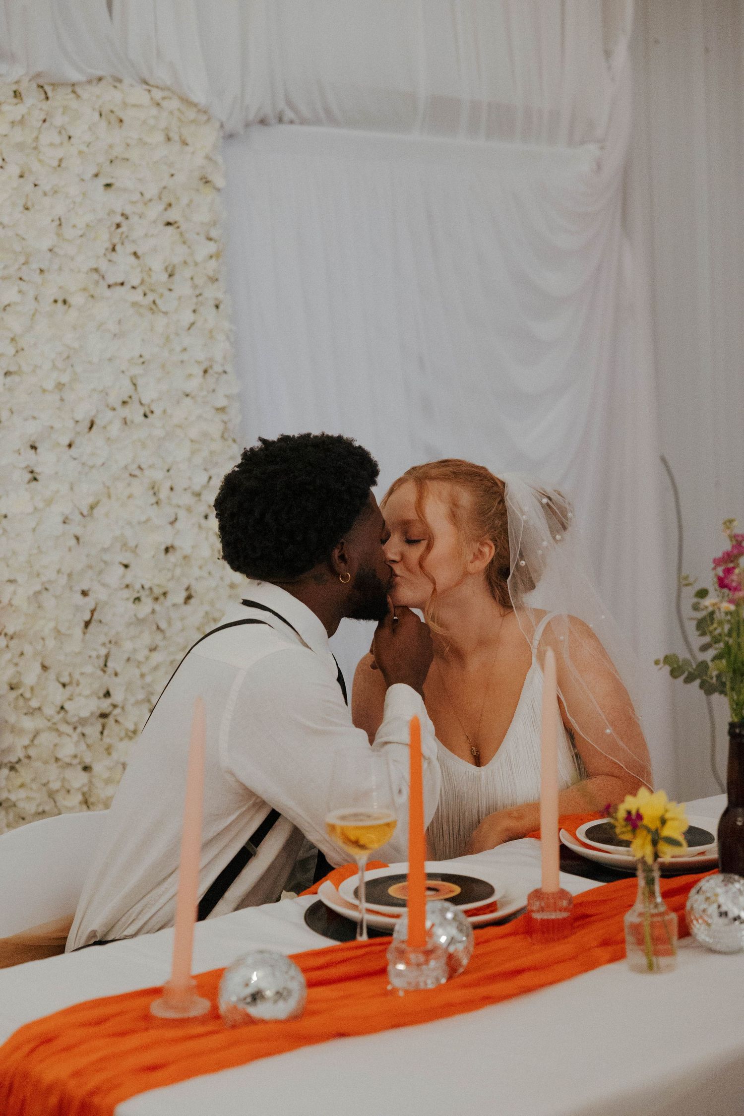 Wedding reception table decorated with orange accents and white floral backdrop.