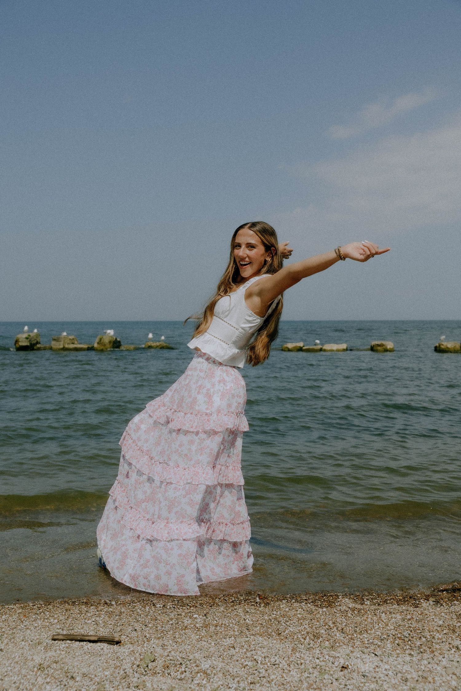 A person in a flowing pink dress dances joyfully on a beach with the ocean and rocks in the background.