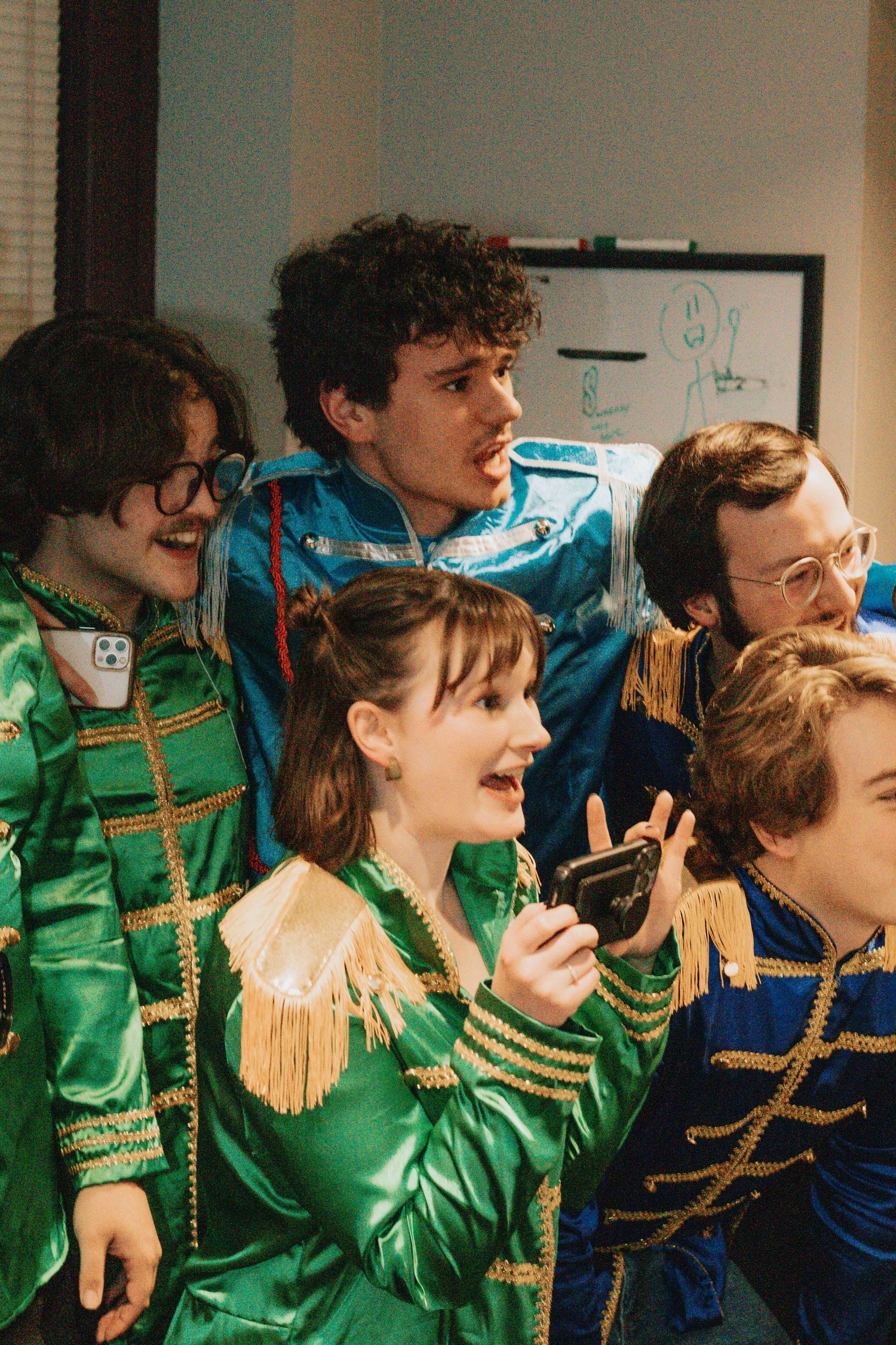 Performers in green and blue military-style uniforms with gold trim sing together on stage.